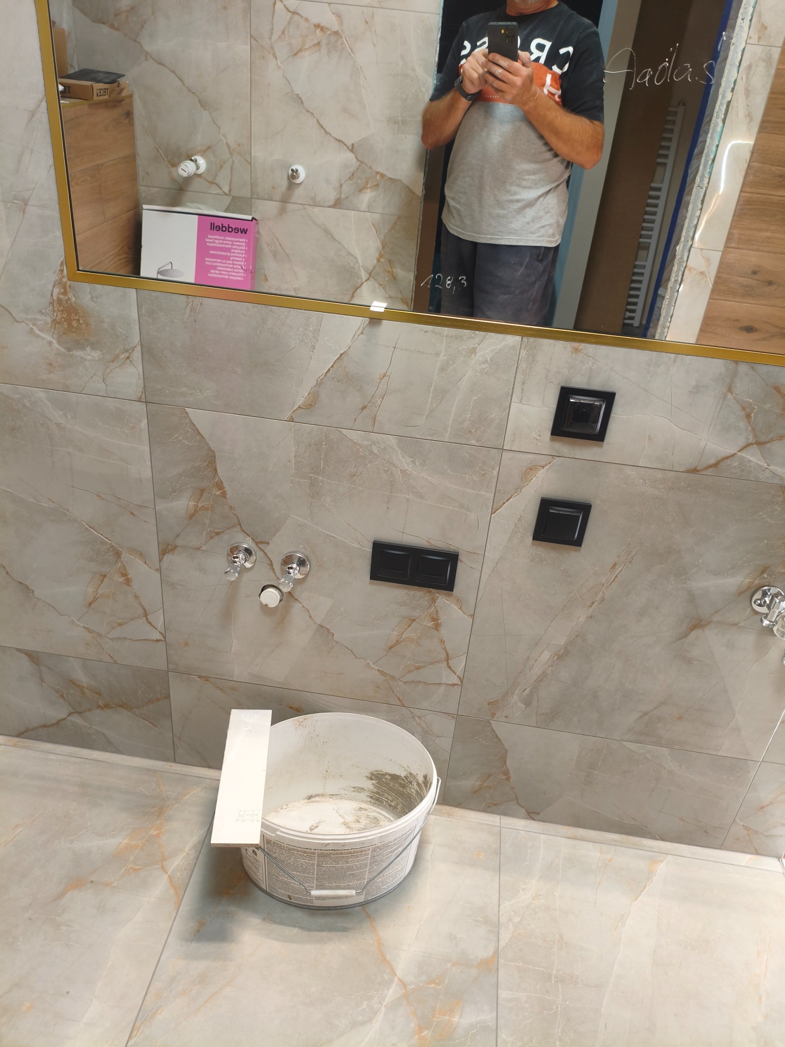 Wykończona łazienka z marmurowymi płytkami, widoczne detale: bateria, włączniki, lustro z odbiciem osoby i wiadrem. Jasne, ciepłe kolory.