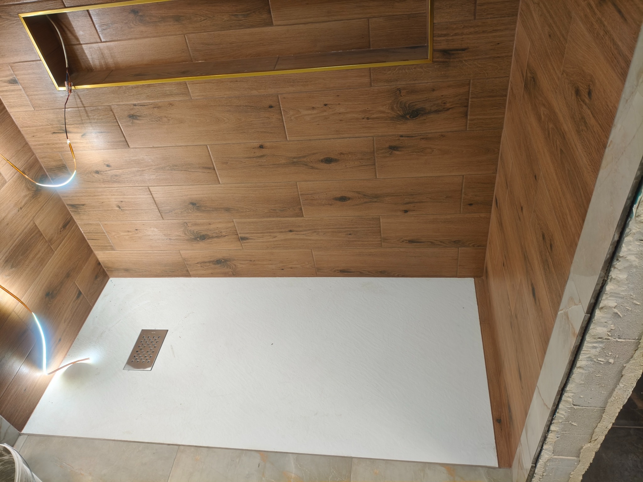 Wykończona kabina prysznicowa z drewnopodobnymi płytkami na ścianach i białym brodzikiem z odpływem liniowym. Widoczne oświetlenie LED i fragment surowej ściany.