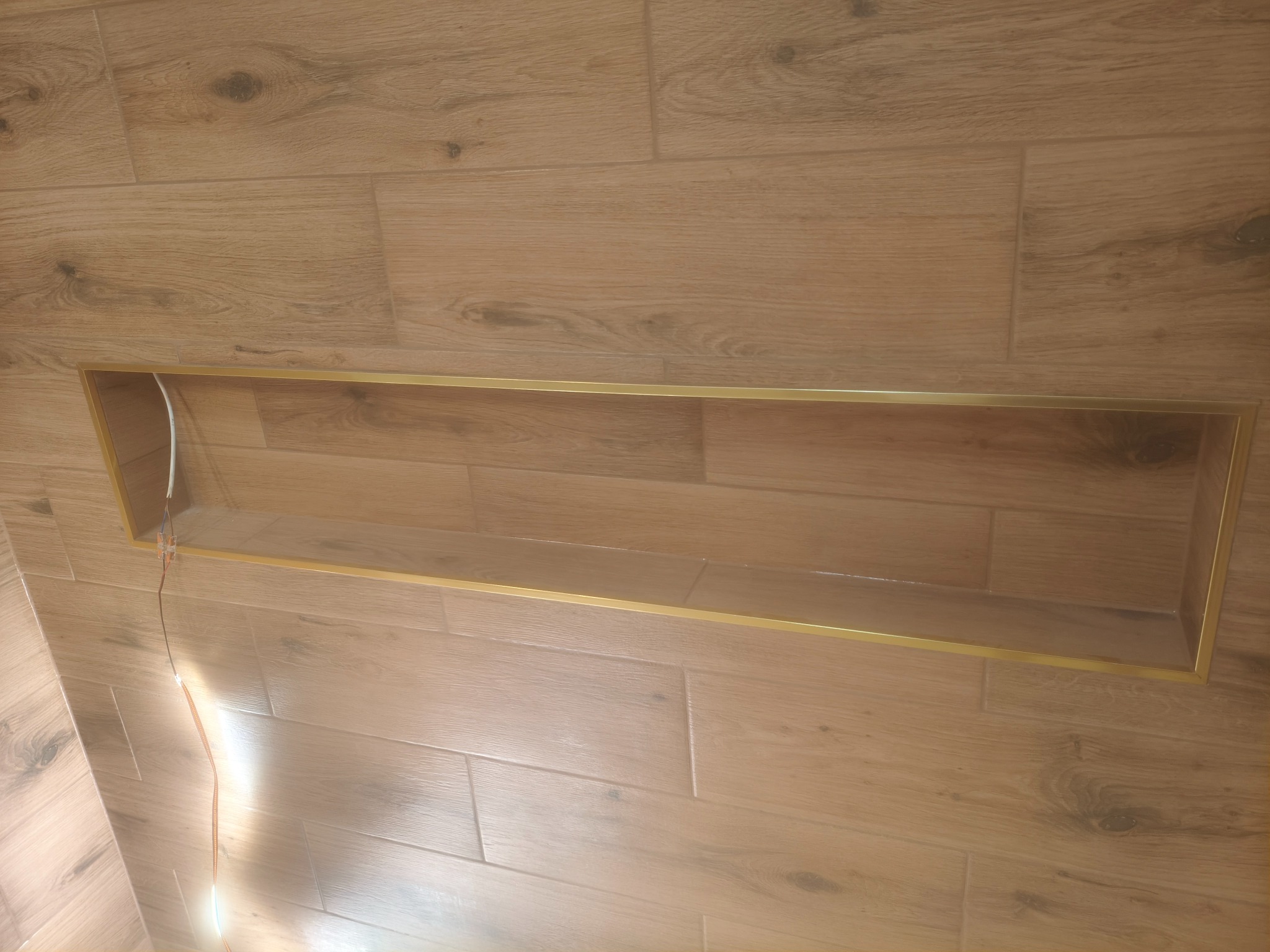 Wykończona wnęka z oświetleniem LED w ścianie wyłożonej płytkami imitującymi drewno, z elegancką, złotą ramką. Widoczne przewody elektryczne.
