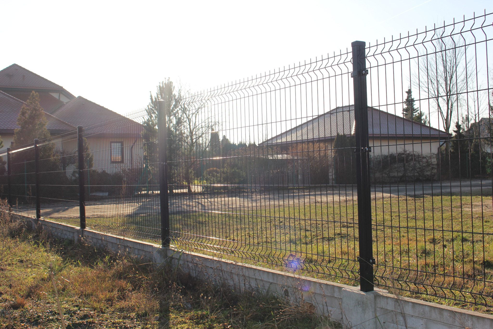 Nowoczesne ogrodzenie metalowe z podmurówką betonową, widok z zewnątrz posesji. Ciemna, siatkowa konstrukcja na tle domów jednorodzinnych w słoneczny dzień.