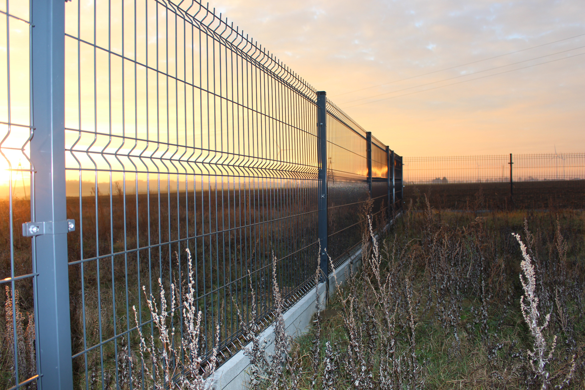 Długi, metalowy płot panelowy w kolorze szarym, z widocznymi elementami montażowymi, na tle zachodzącego słońca i łąki. W tle widoczny drugi płot.