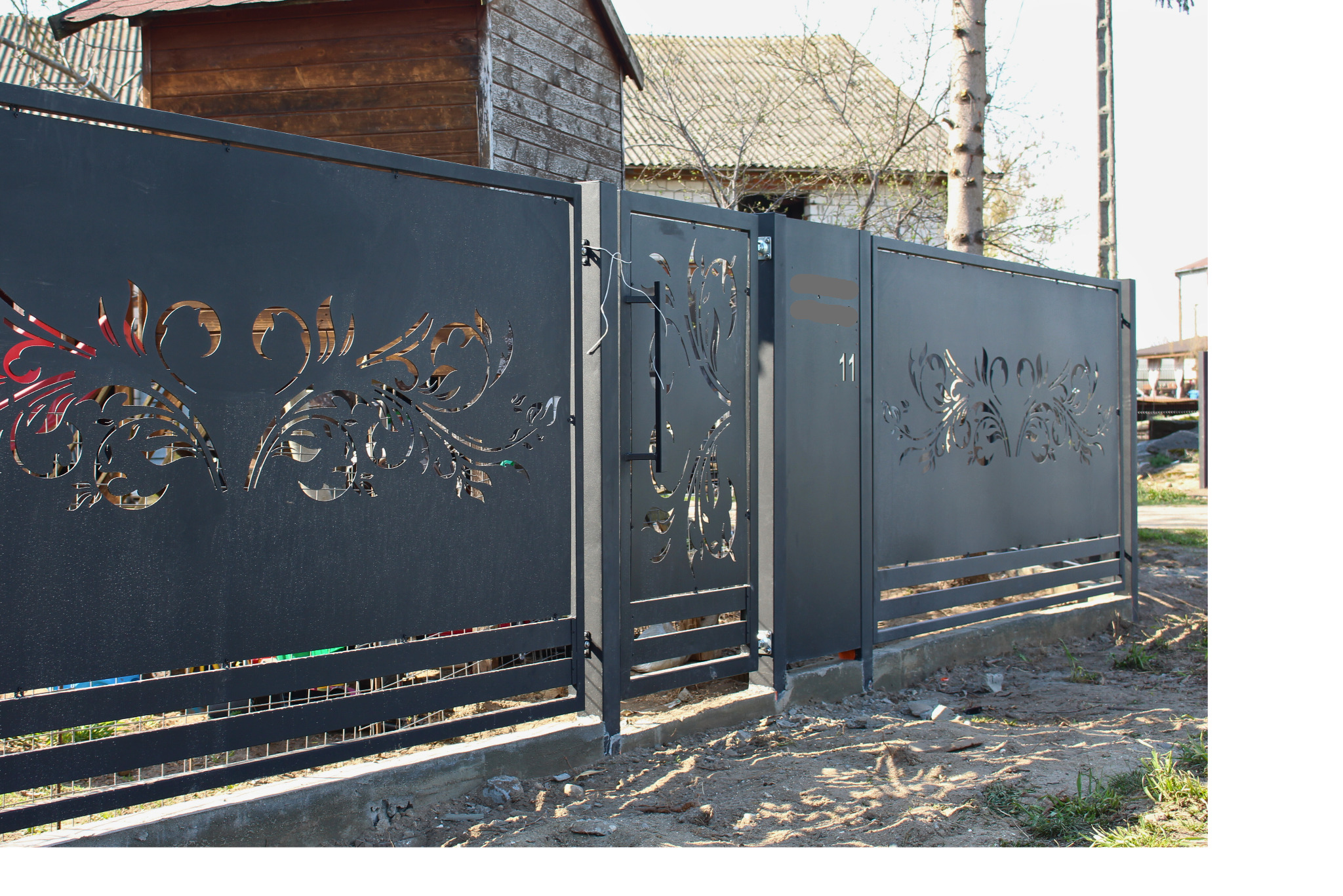 Nowoczesne, metalowe ogrodzenie z wycinanym laserowo, florystycznym wzorem, w kolorze grafitowym. Widoczna brama wejściowa i fragment posesji w tle.