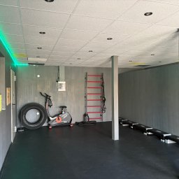 RuszDoope.pl Aktywny Wypoczynek Justyna Snarska - Nowoczesna siłownia w Radomiu z rowerem stacjonarnym, oponą, drabinką gimnastyczną i stepami. Minimalistyczny design, szare ściany, czarna podłoga i zielone oświetlenie LED.