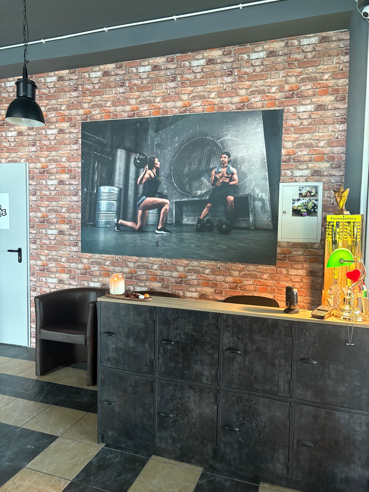 Wnętrze siłowni: ceglana ściana z plakatem ćwiczących osób, recepcja z dekoracjami i krzesłem. Czarno-biała podłoga w paski. Klimatyczne oświetlenie.