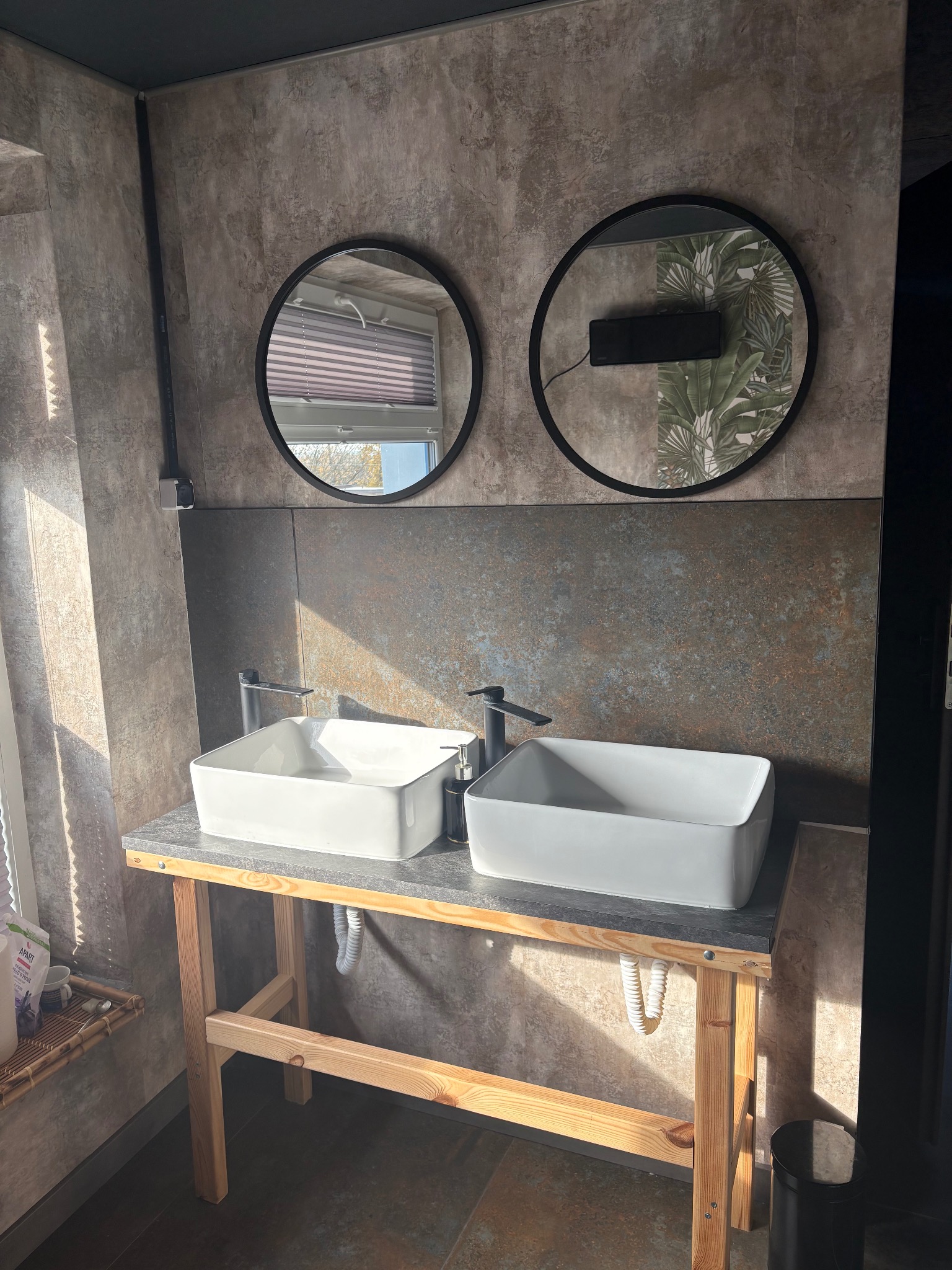 Dwie białe umywalki nablatowe na drewnianym stelażu, okrągłe lustra z czarną ramą na tle ściany z efektem betonu. Nowoczesna aranżacja łazienki.