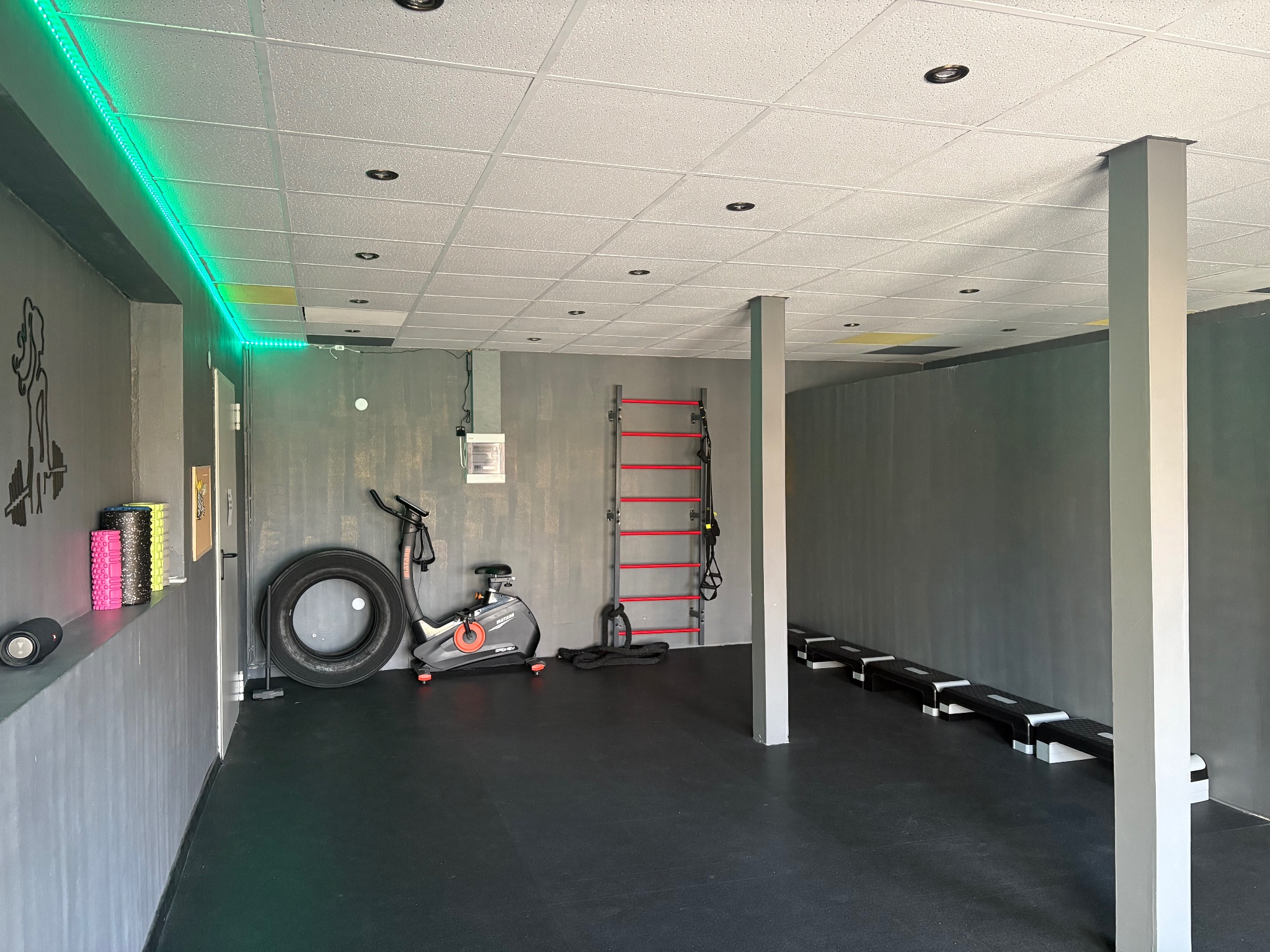 Nowoczesna siłownia w Radomiu z rowerem stacjonarnym, oponą, drabinką gimnastyczną i stepami. Minimalistyczny design, szare ściany, czarna podłoga i zielone oświetlenie LED.