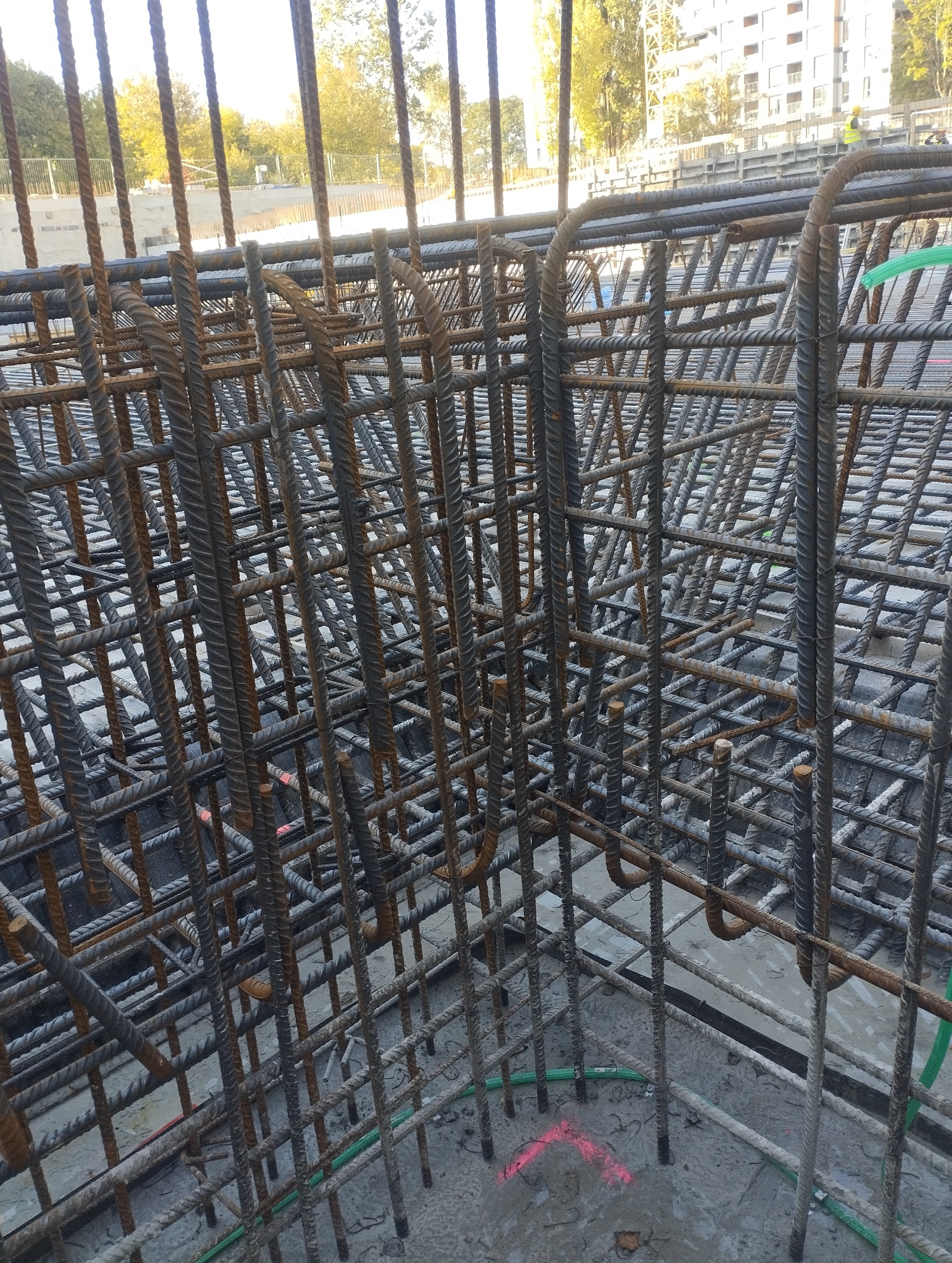 Skomplikowana siatka zbrojeniowa z prętów stalowych, widok z góry, z elementami zielonej rurki i różowego oznaczenia na szarym betonie. W tle budynek i drzewa.