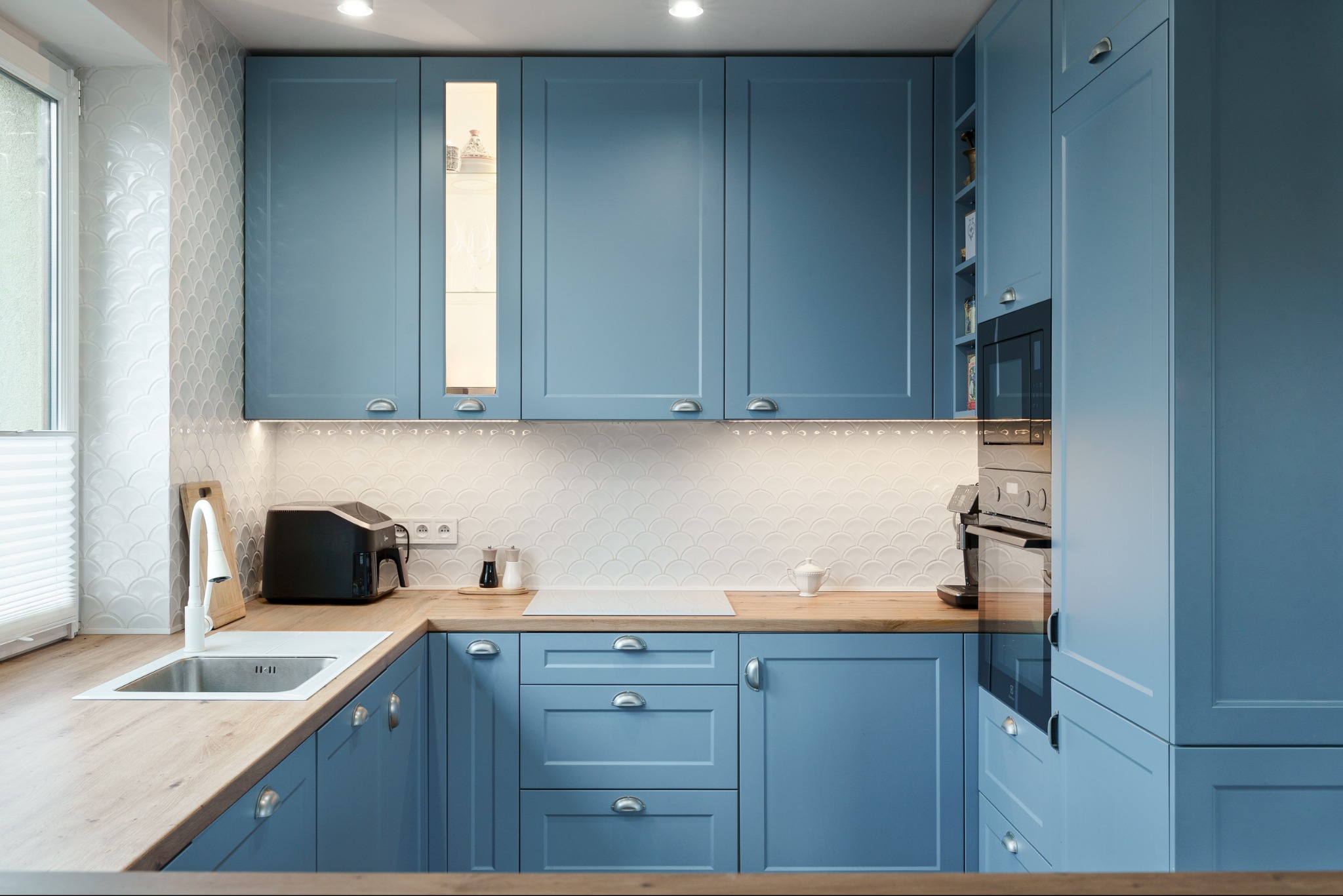Błękitna kuchnia z drewnianym blatem i białymi płytkami w kształcie rybiej łuski. Oświetlenie podszafkowe podkreśla detale. Nowoczesny design z elementami klasyki.