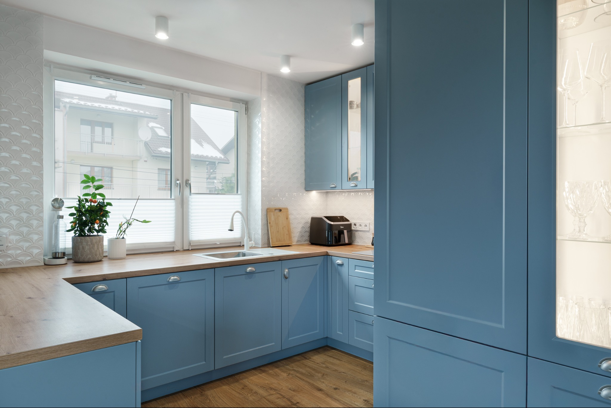 Błękitna kuchnia z drewnianym blatem i podłogą. W tle okno z widokiem na dom. Na blacie roślina w doniczce i akcesoria kuchenne. Nowoczesny design.