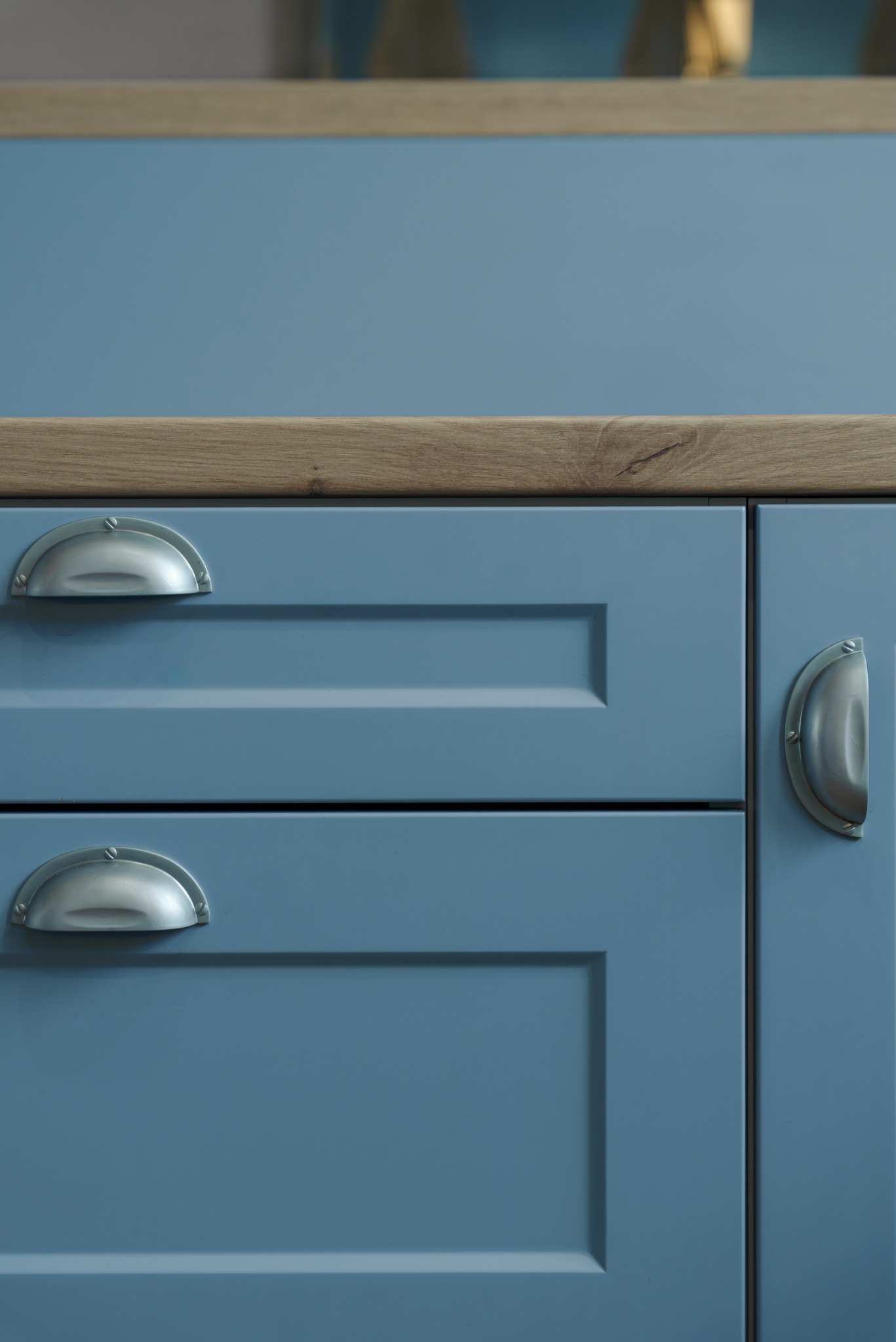 Niebieskie fronty szafek kuchennych z drewnianym blatem i metalowymi uchwytami w kształcie muszelek. Detal wykończenia mebli kuchennych.