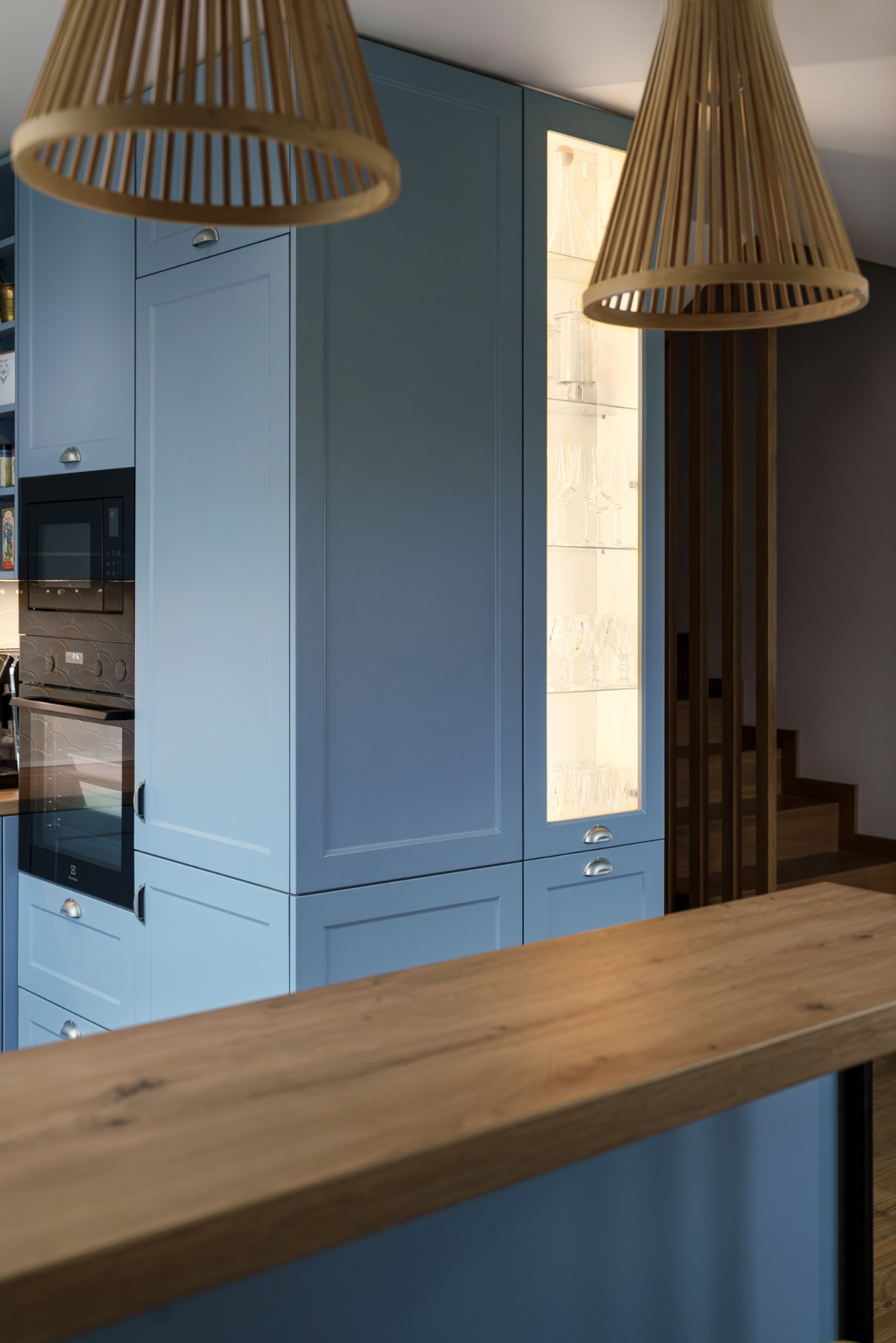 Błękitna zabudowa kuchenna z oświetloną witryną na kieliszki, drewniany blat i lampy z abażurami z naturalnego materiału. Widoczne schody w tle.