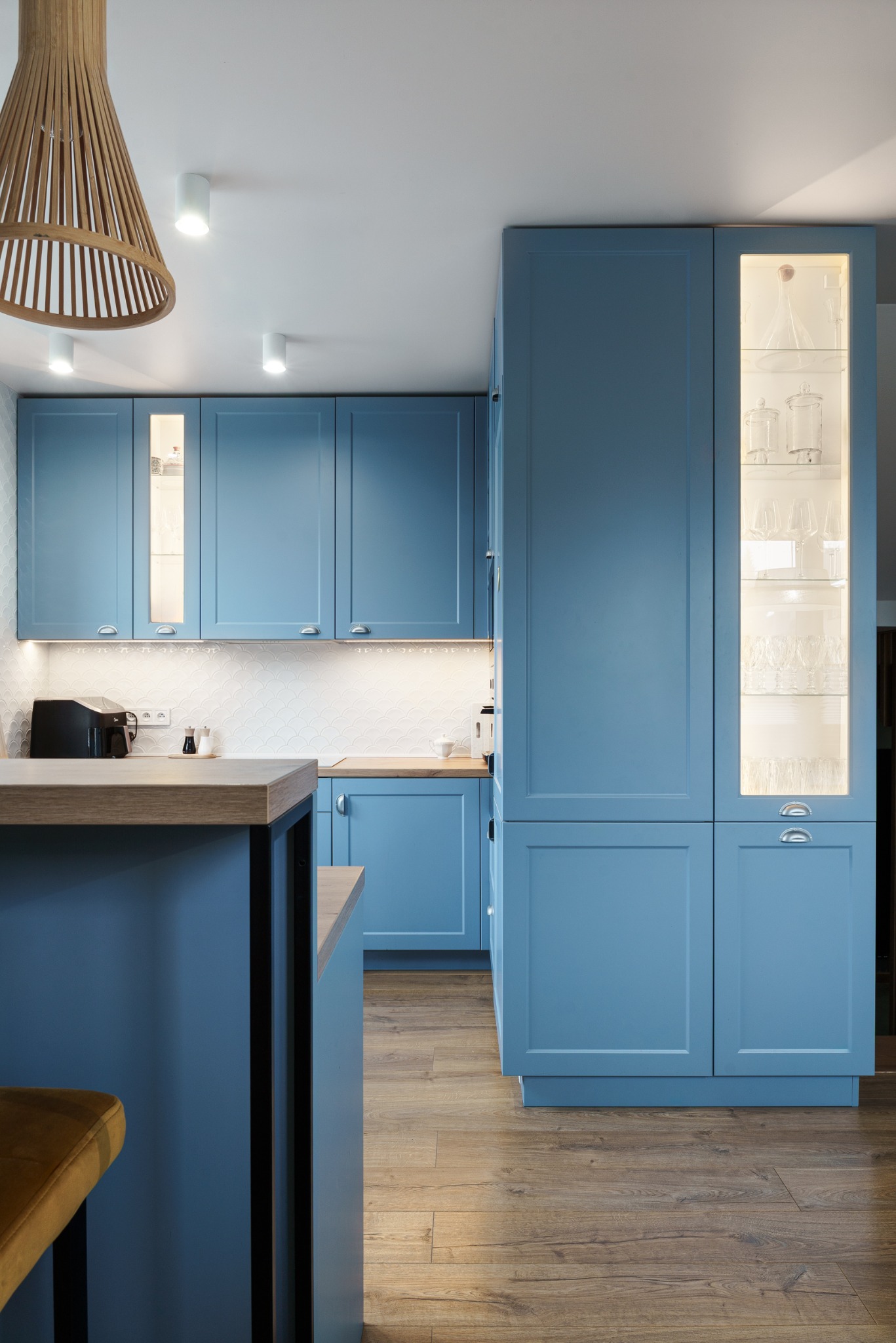 Błękitna zabudowa kuchenna z drewnianym blatem i oświetleniem LED, uzupełniona dekoracyjną lampą wiszącą i drewnianą podłogą. Nowoczesny design.