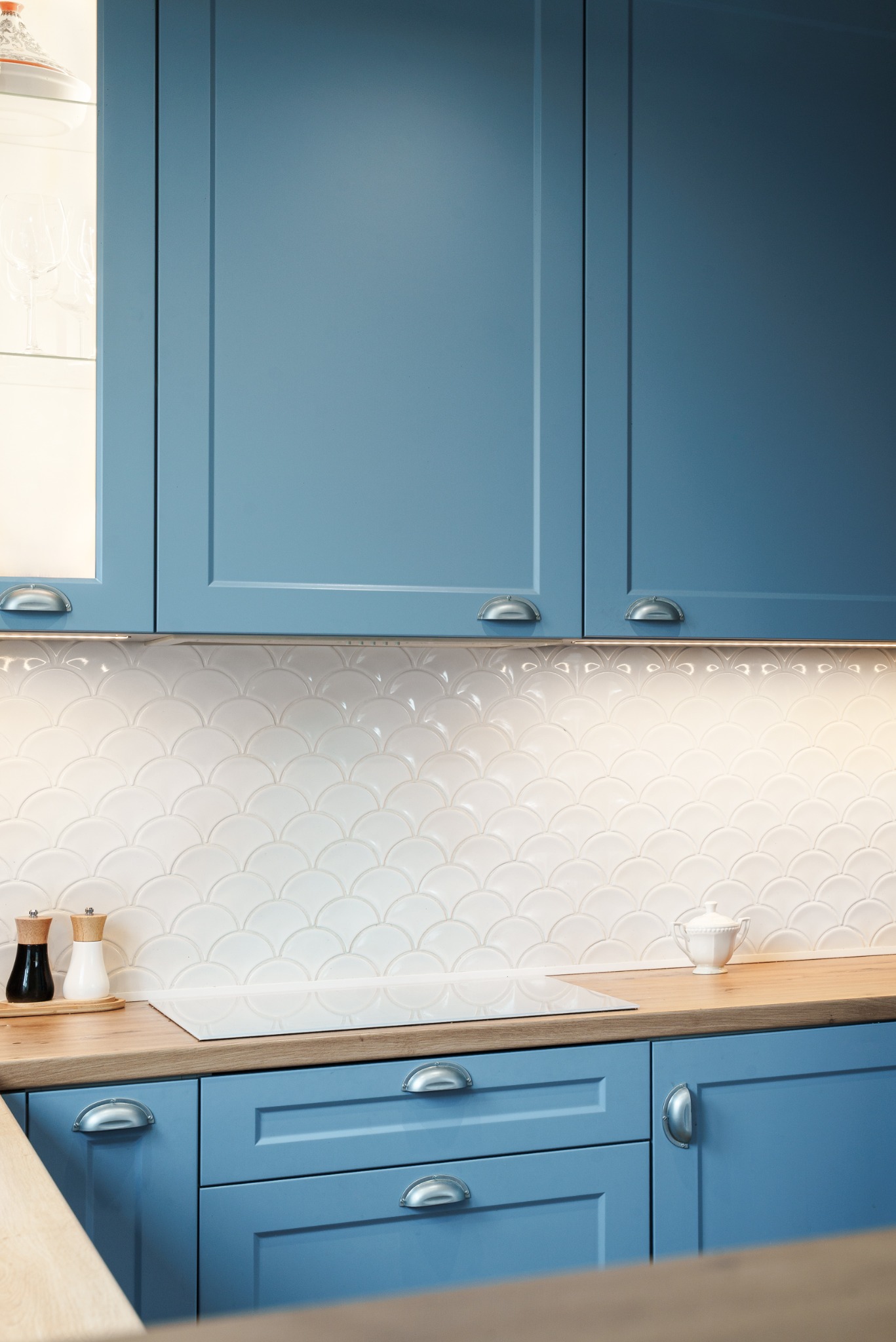 Błękitne szafki kuchenne z frezowanymi frontami i srebrnymi uchwytami typu muszelka. Białe płytki w kształcie rybiej łuski i drewniany blat. Oświetlenie LED podszafkowe.