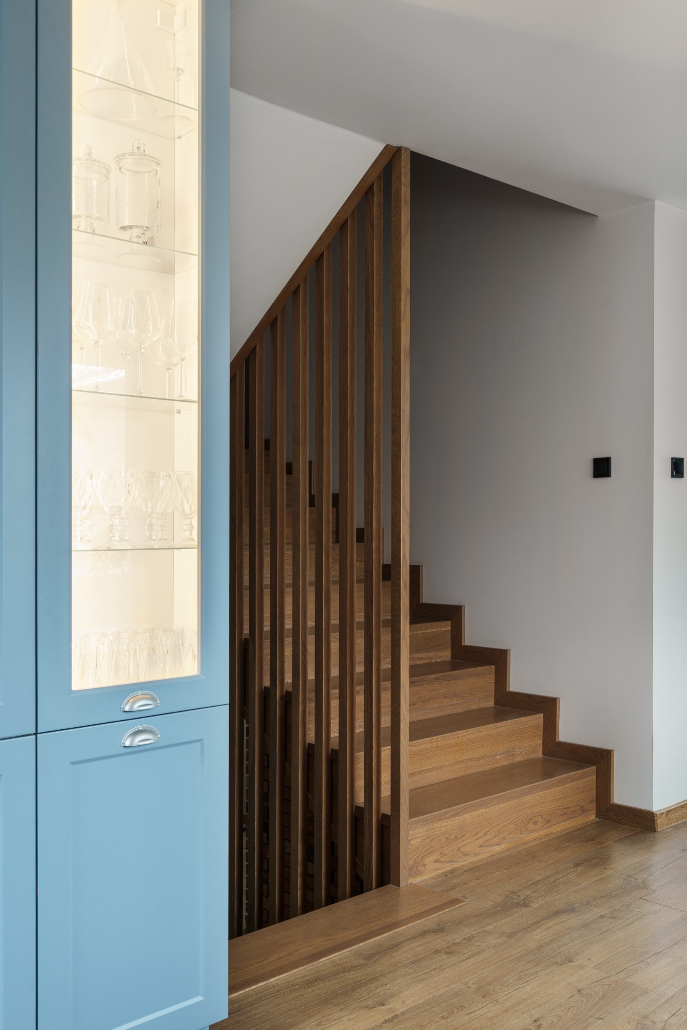 Błękitna witryna z oświetleniem i szklanymi naczyniami obok drewnianych schodów z ażurową balustradą. Minimalistyczne wnętrze z parkietem i czarnymi włącznikami.