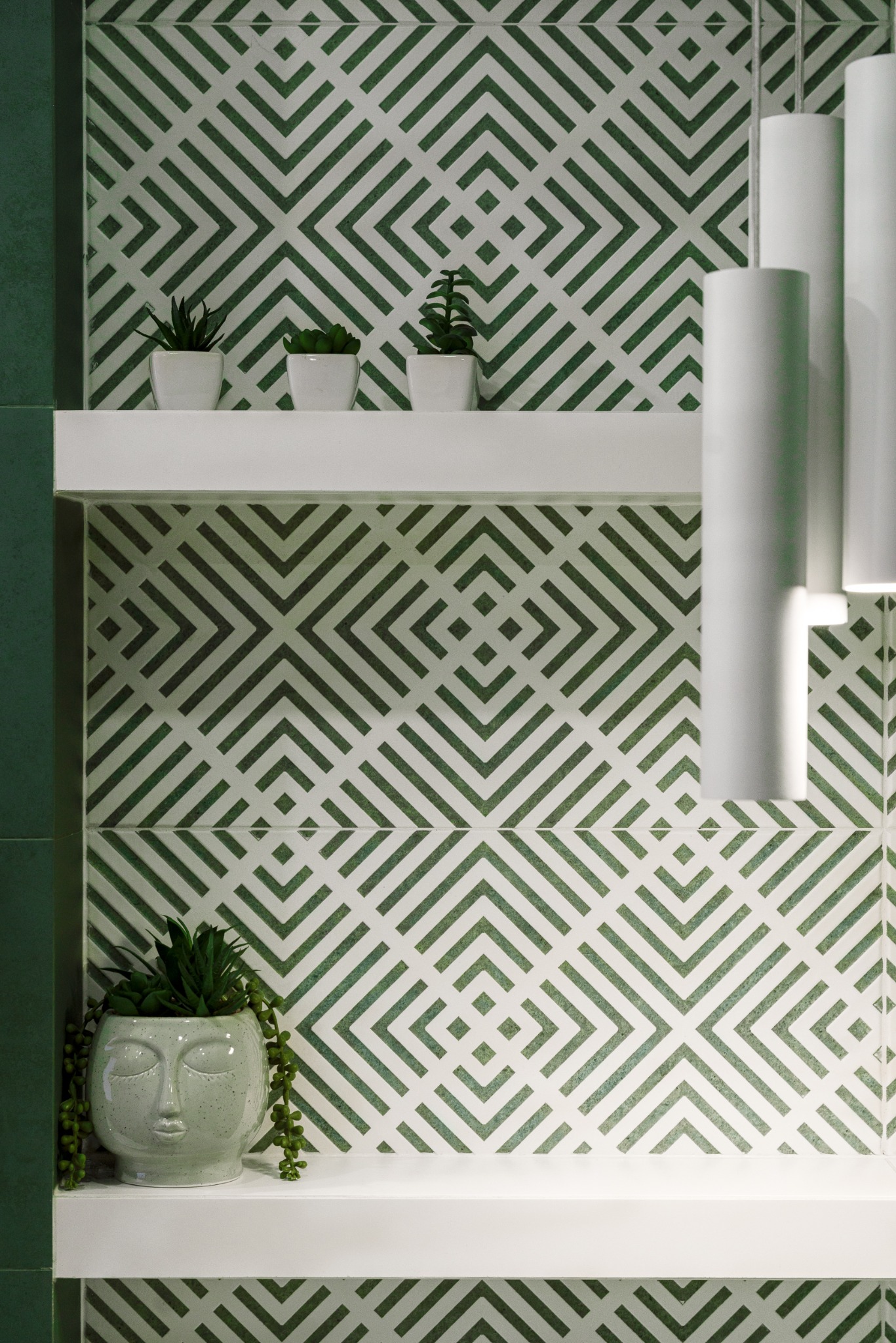 Nowoczesna łazienka z zielono-białymi płytkami w geometryczny wzór, białe półki z roślinami i cylindryczne lampy wiszące. Minimalistyczny design.