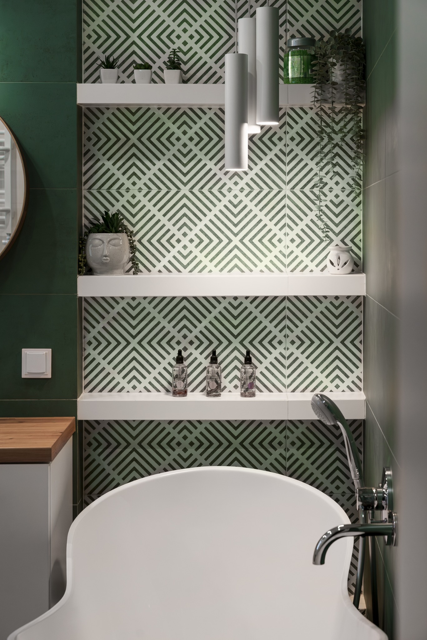 Nowoczesna łazienka z wanną wolnostojącą, zielonymi płytkami w geometryczny wzór i białymi półkami z dekoracjami. Minimalistyczne oświetlenie.