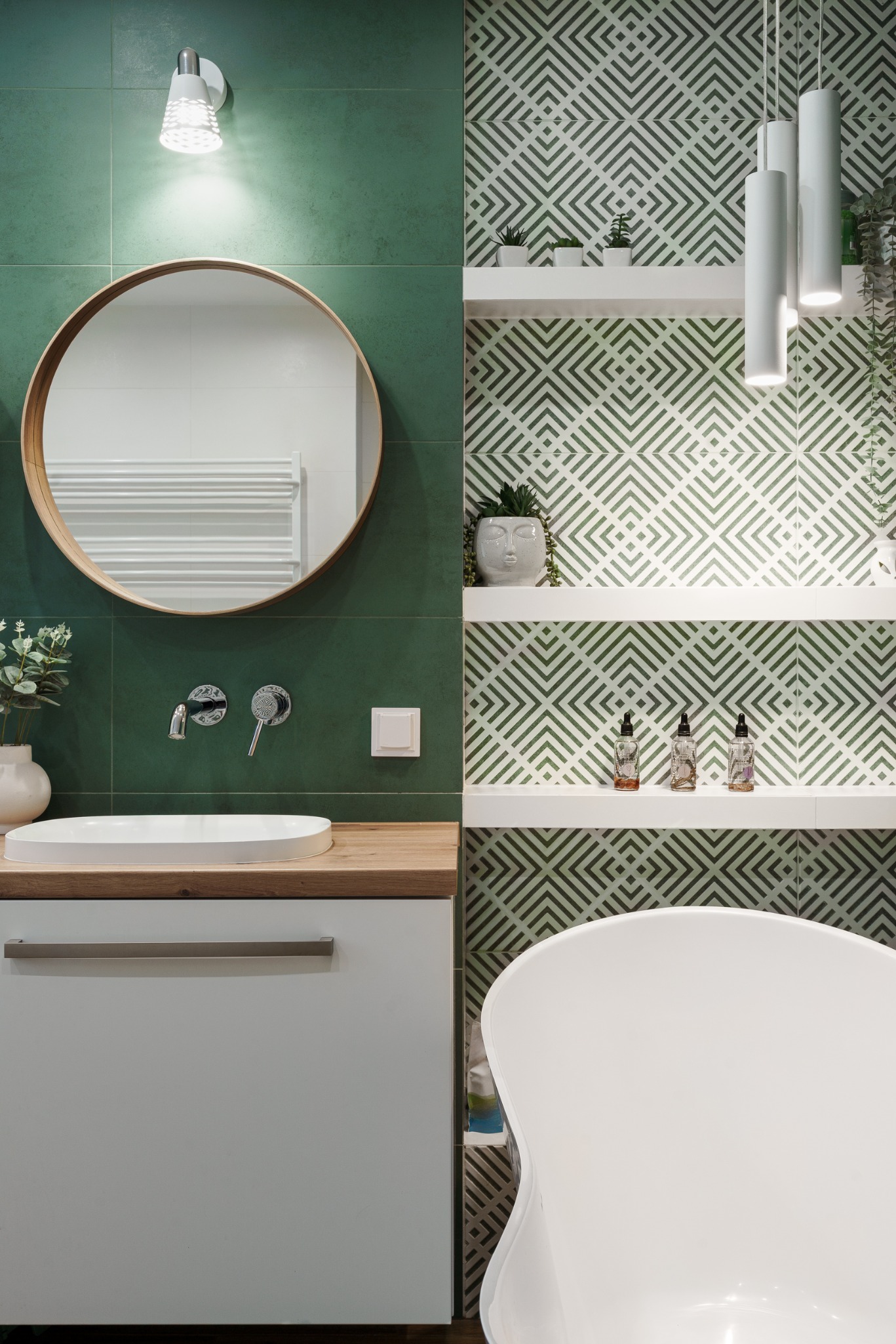 Nowoczesna łazienka z zielonymi płytkami, okrągłym lustrem w drewnianej ramie, białą umywalką nablatową i geometrycznym wzorem na ścianie.