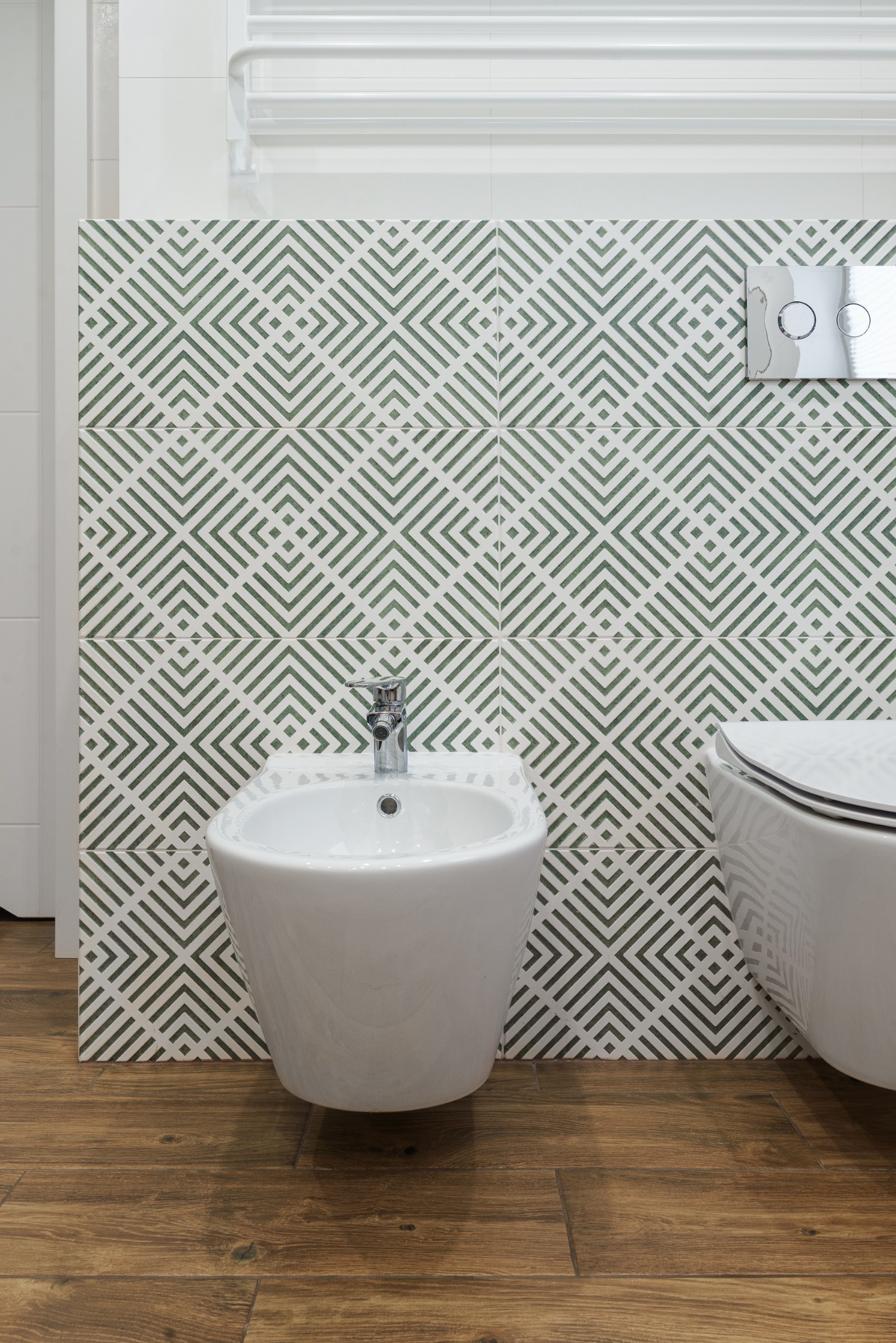 Nowoczesna łazienka z bidetem i toaletą, zielono-białe płytki z geometrycznym wzorem, drewnopodobna podłoga. Minimalistyczny design, chromowana armatura.