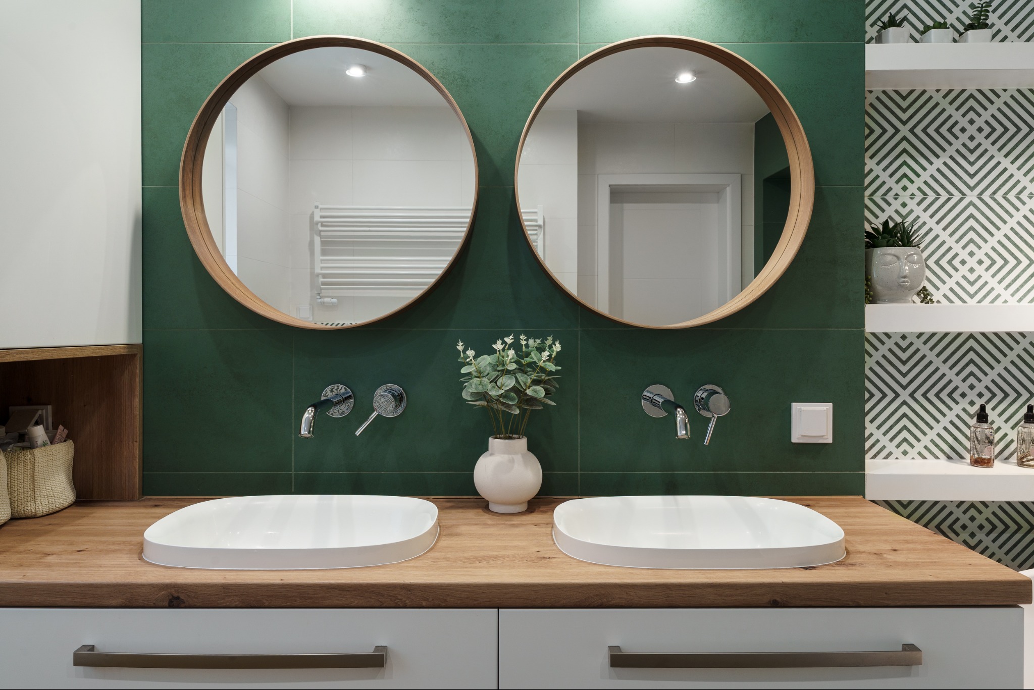 Nowoczesna łazienka z dwoma okrągłymi lustrami nad podwójną umywalką na drewnianym blacie i zielonymi płytkami. Minimalistyczny design z roślinami i geometrycznym wzorem.
