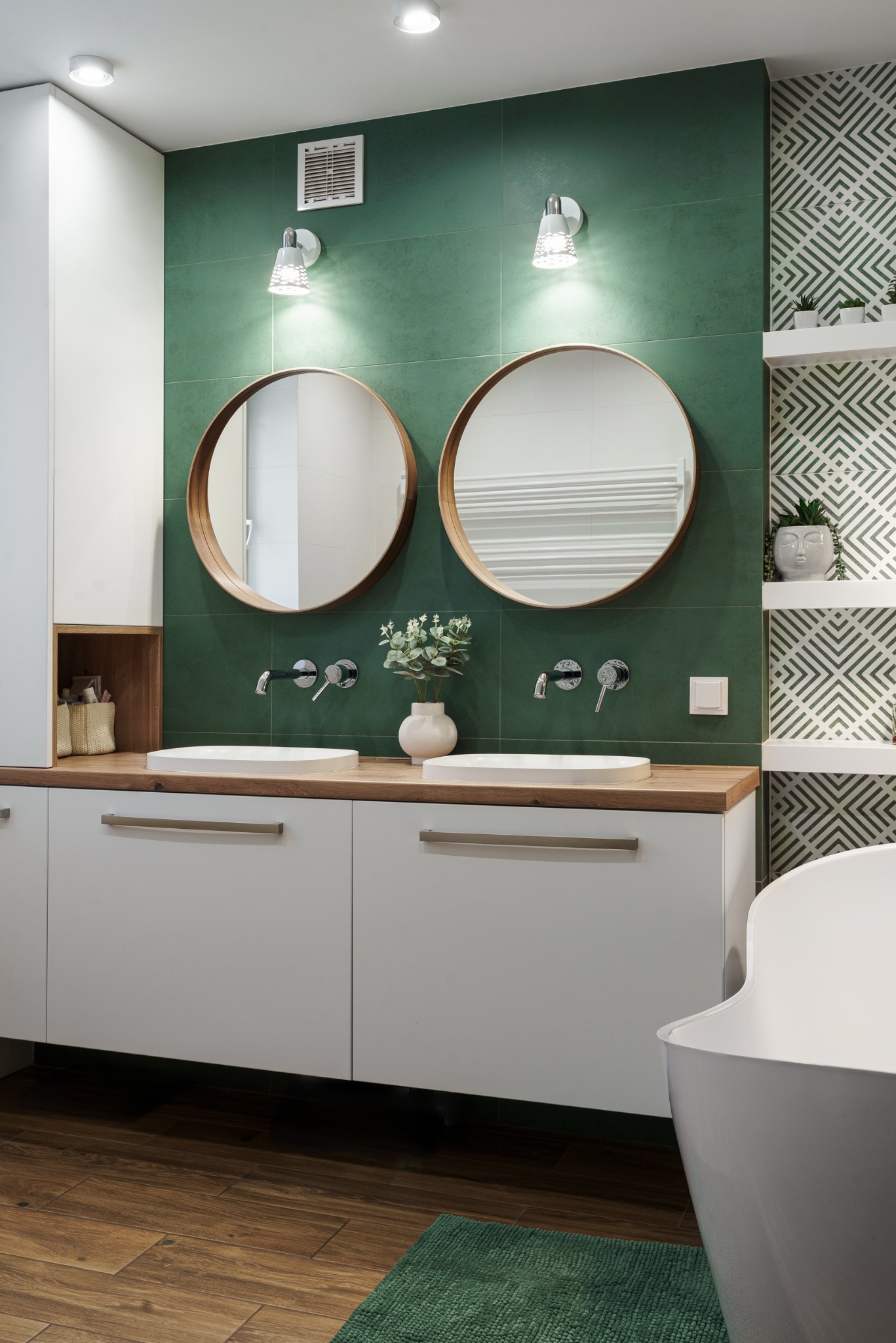 Nowoczesna łazienka z dwoma okrągłymi lustrami nad umywalkami, zielonymi płytkami i drewnianym blatem. Widoczna wanna i półki z dekoracjami.