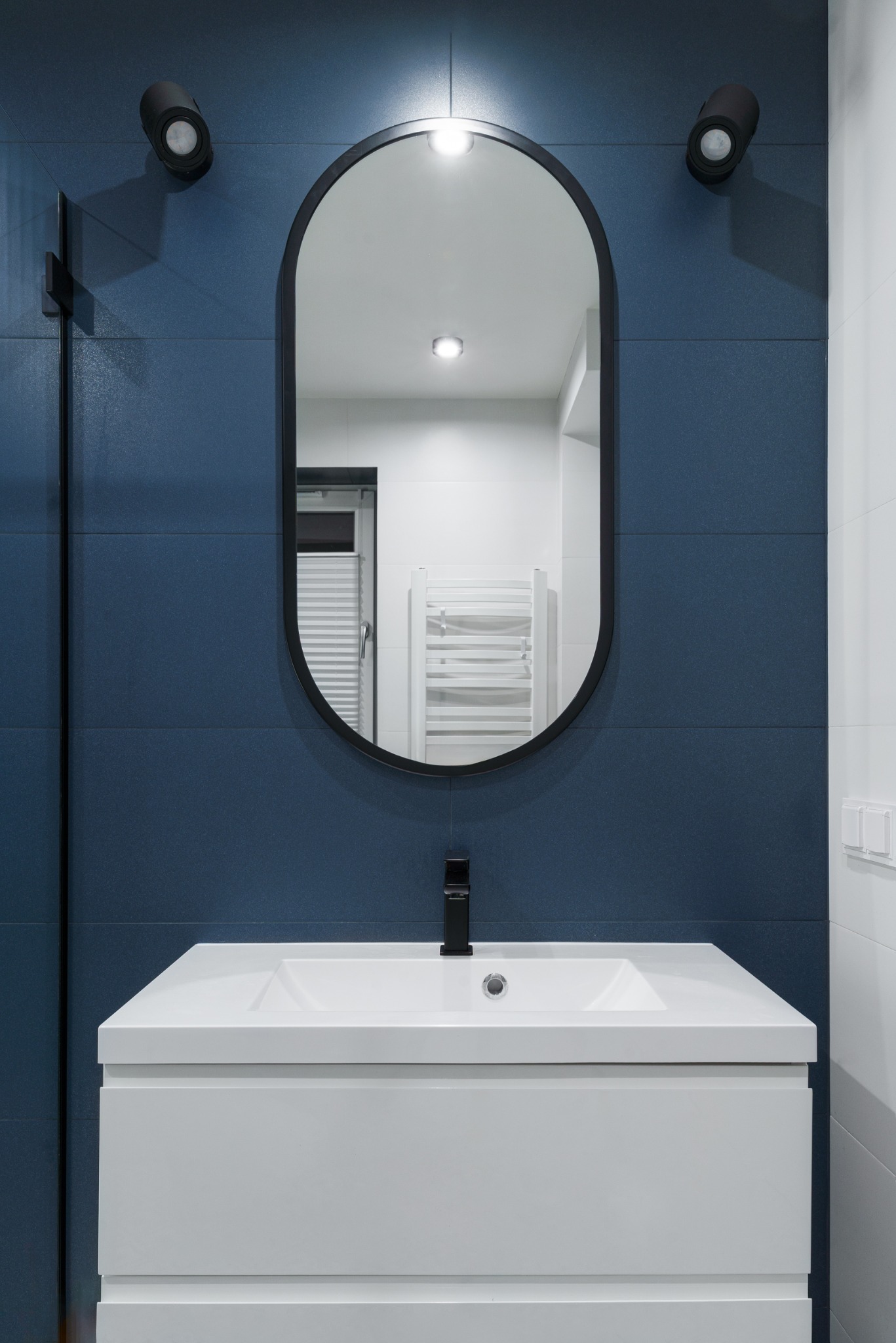 Nowoczesna łazienka z umywalką na szafce, owalnym lustrem w czarnej ramie i ciemnoniebieskimi płytkami. Minimalistyczne oświetlenie punktowe.