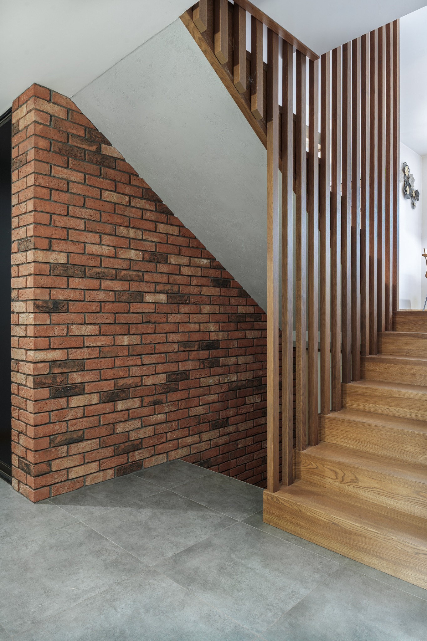 Ceglana ściana z drewnianą balustradą schodów w nowoczesnym wnętrzu. Szare płytki podłogowe i minimalistyczny design.