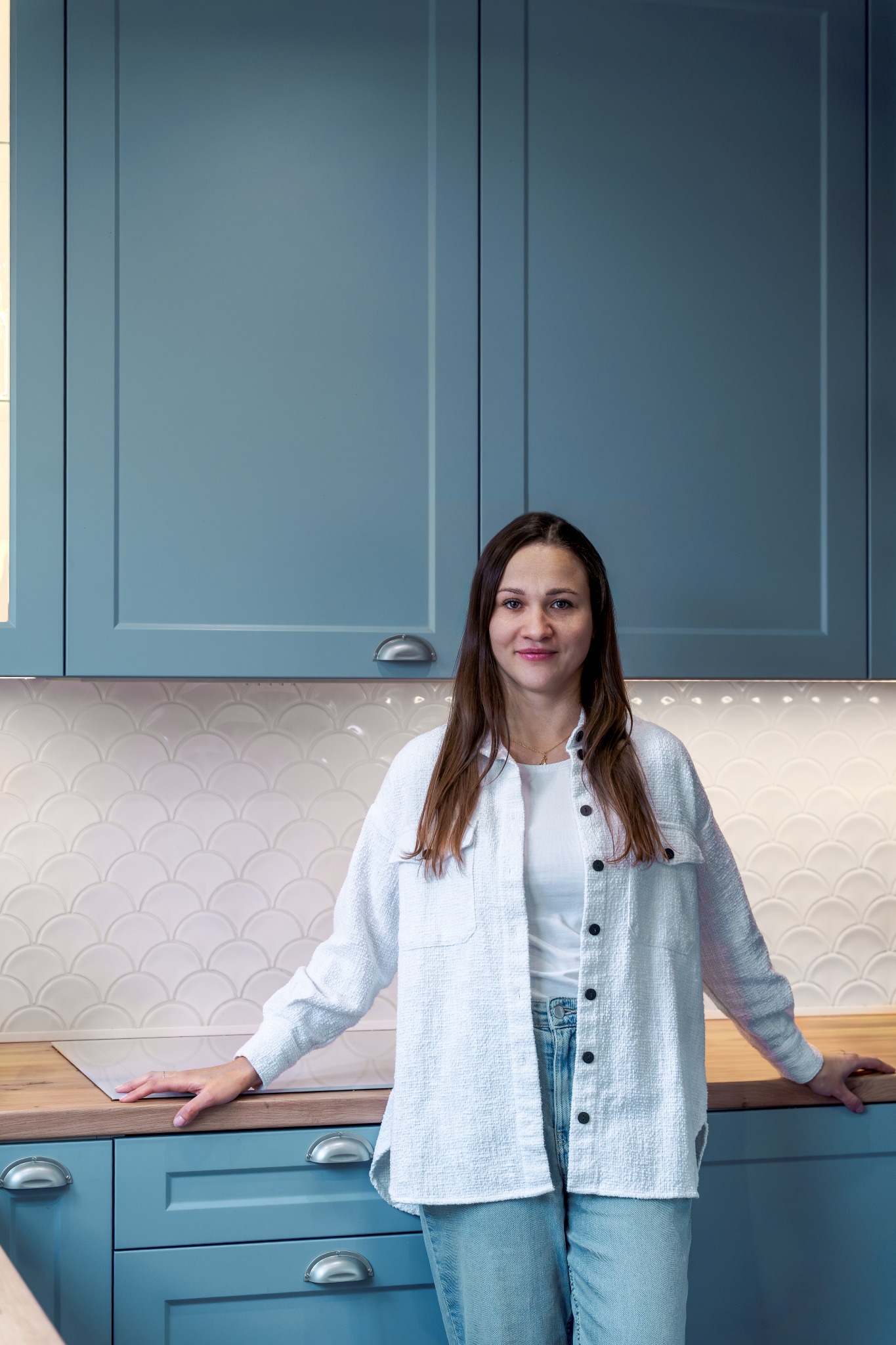 Kobieta w kuchni z błękitnymi szafkami i blatem z drewna. Białe płytki w kształcie rybiej łuski. Uśmiechnięta kobieta opiera się o blat.