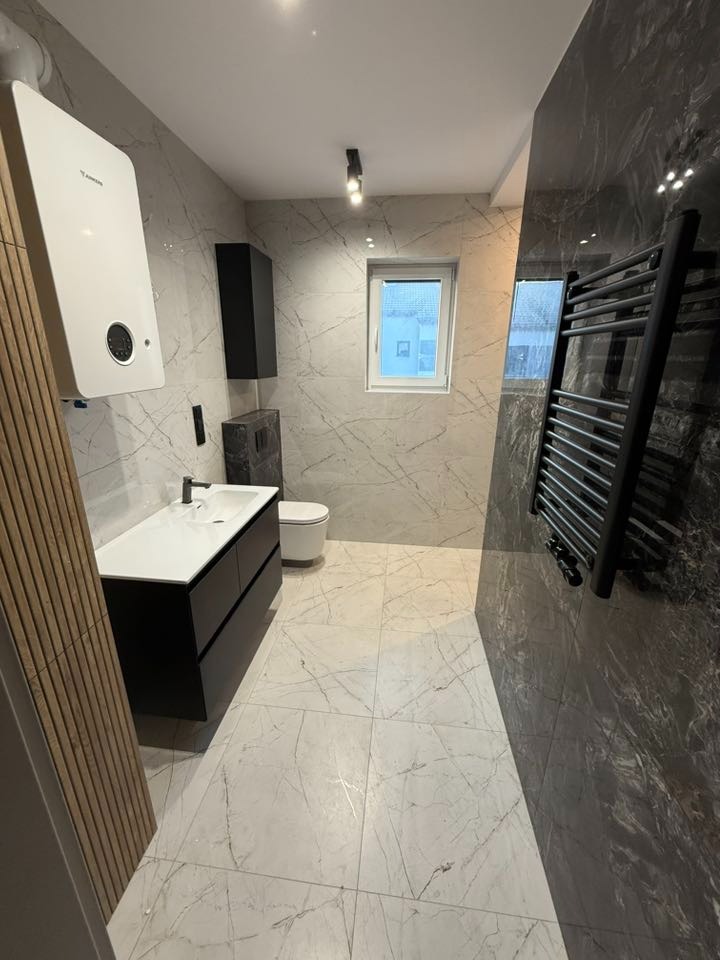 Nowoczesna łazienka z marmurowymi płytkami, czarną armaturą i grzejnikiem drabinkowym. Widoczny bojler Junkers i szafka pod umywalką. Minimalistyczny design.