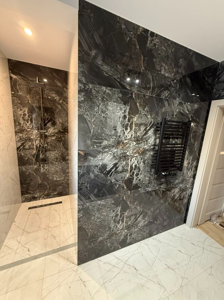 Elegancka łazienka z kabiną typu walk-in, wykończona płytkami imitującymi marmur w odcieniach bieli i czerni. Widoczny grzejnik drabinkowy i odpływ liniowy.