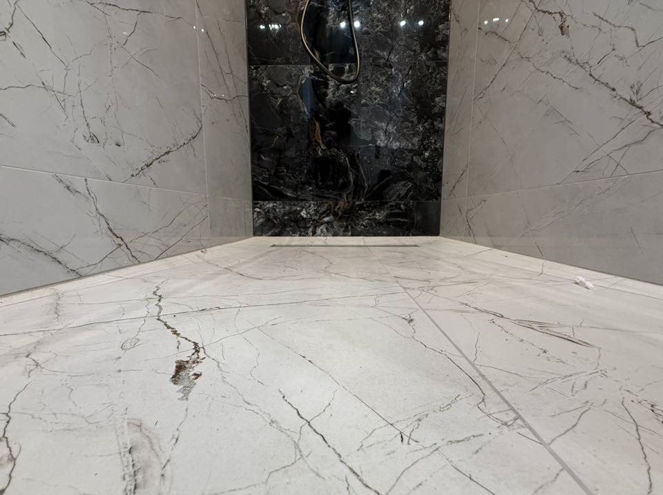 Wykończona kabina prysznicowa z białymi płytkami imitującymi marmur na ścianach i podłodze, oraz czarną, błyszczącą ścianą akcentującą. Widoczny odpływ liniowy.