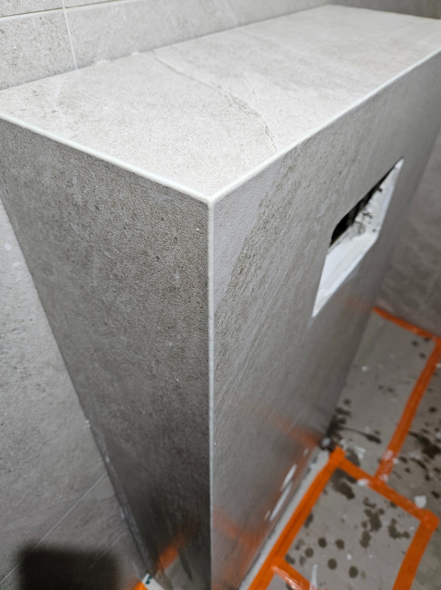 Obudowa z płytek w łazience, widoczny otwór instalacyjny i pomarańczowe linie wyznaczające obszar robót. Szare płytki z delikatną fakturą, fuga w jasnym odcieniu.