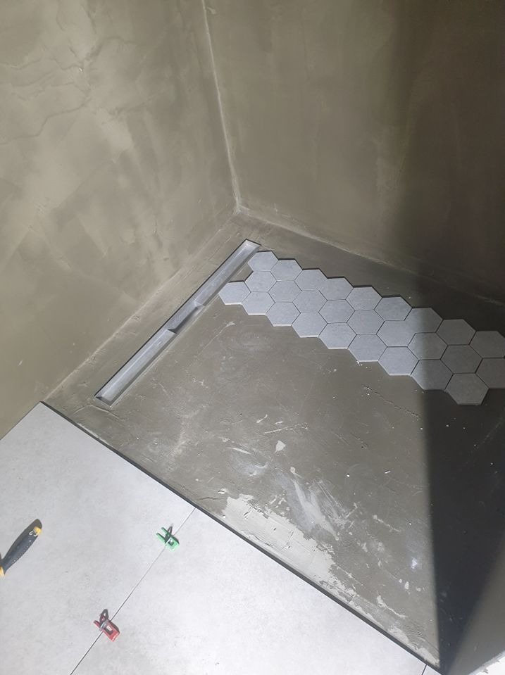 Układanie szarych, heksagonalnych płytek w strefie prysznicowej z odpływem liniowym. Widoczne narzędzia glazurnicze i surowe ściany przygotowane do dalszych prac.