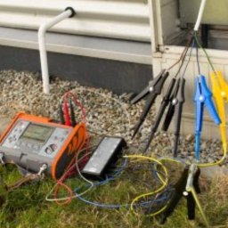 SEBASTIAN SZYMANOWSKI X-SELL - Pomiar rezystancji uziomu: miernik, przewody z krokodylkami wpięte w instalację elektryczną na zewnątrz budynku. Prace pomiarowe w toku, widoczne elementy uziomu.