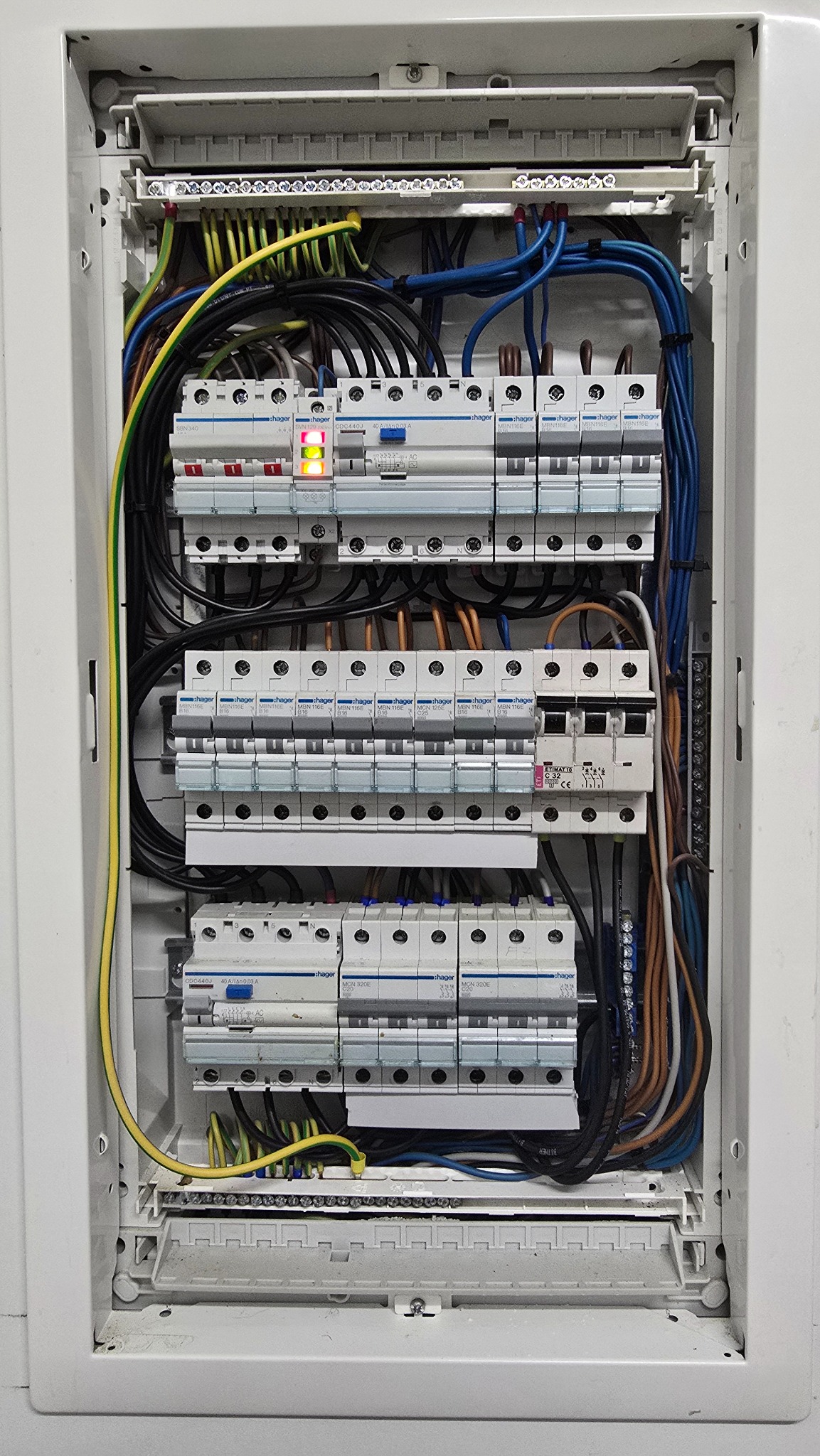 Rozdzielnica elektryczna Hager z przewodami w kolorach: żółto-zielonym, niebieskim i czarnym. Widoczne bezpieczniki i listwy zaciskowe. Jeden z bezpieczników sygnalizuje awarię.