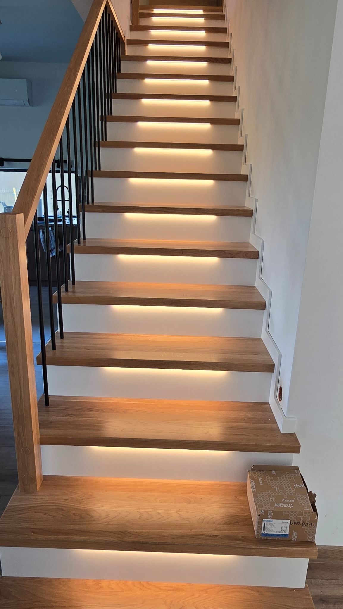 Schody drewniane z podświetleniem LED, białe stopnice, czarna balustrada. Na dole karton z elementami elektrycznymi. Ciepłe światło podkreśla nowoczesny design.