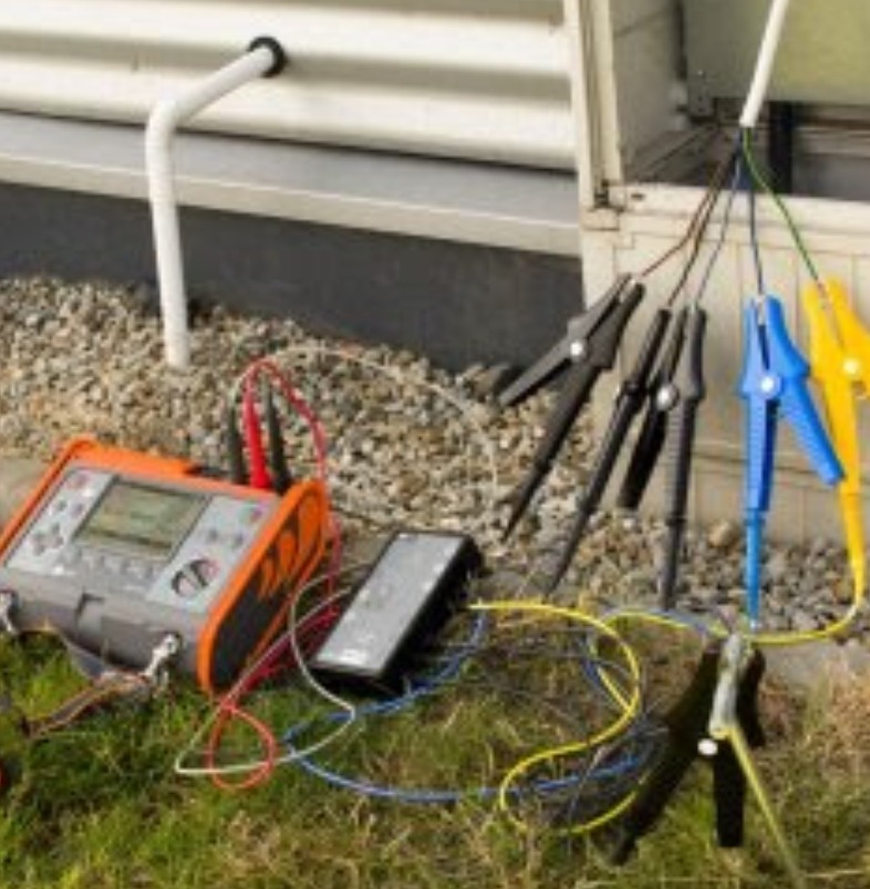 Pomiar rezystancji uziomu: miernik, przewody z krokodylkami wpięte w instalację elektryczną na zewnątrz budynku. Prace pomiarowe w toku, widoczne elementy uziomu.