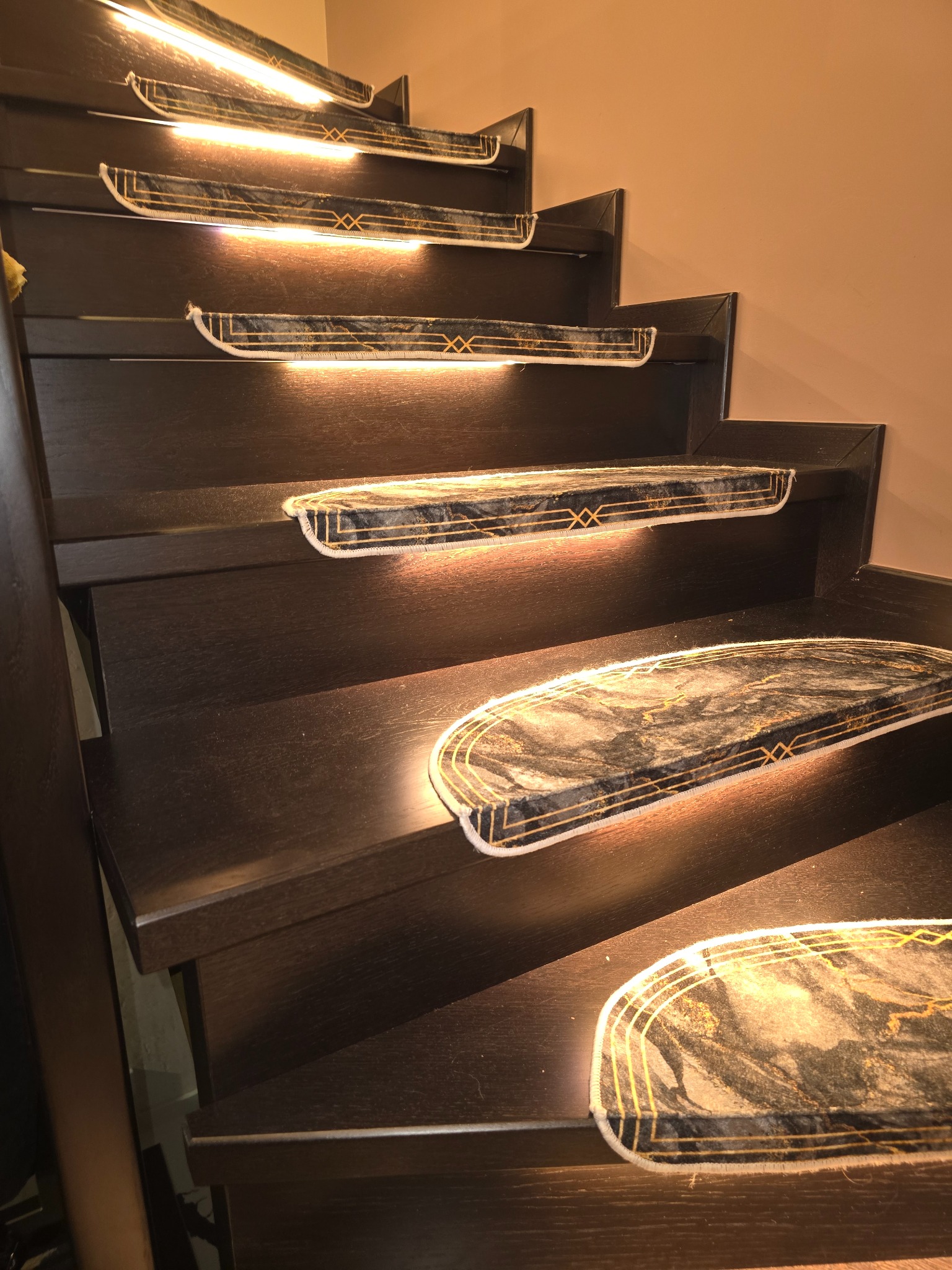 Oświetlone schody z ciemnego drewna z nakładkami w stylu art deco. Ciepłe światło podkreśla wzór i dodaje elegancji wnętrzu. Ujęcie z dołu, ukazujące perspektywę.