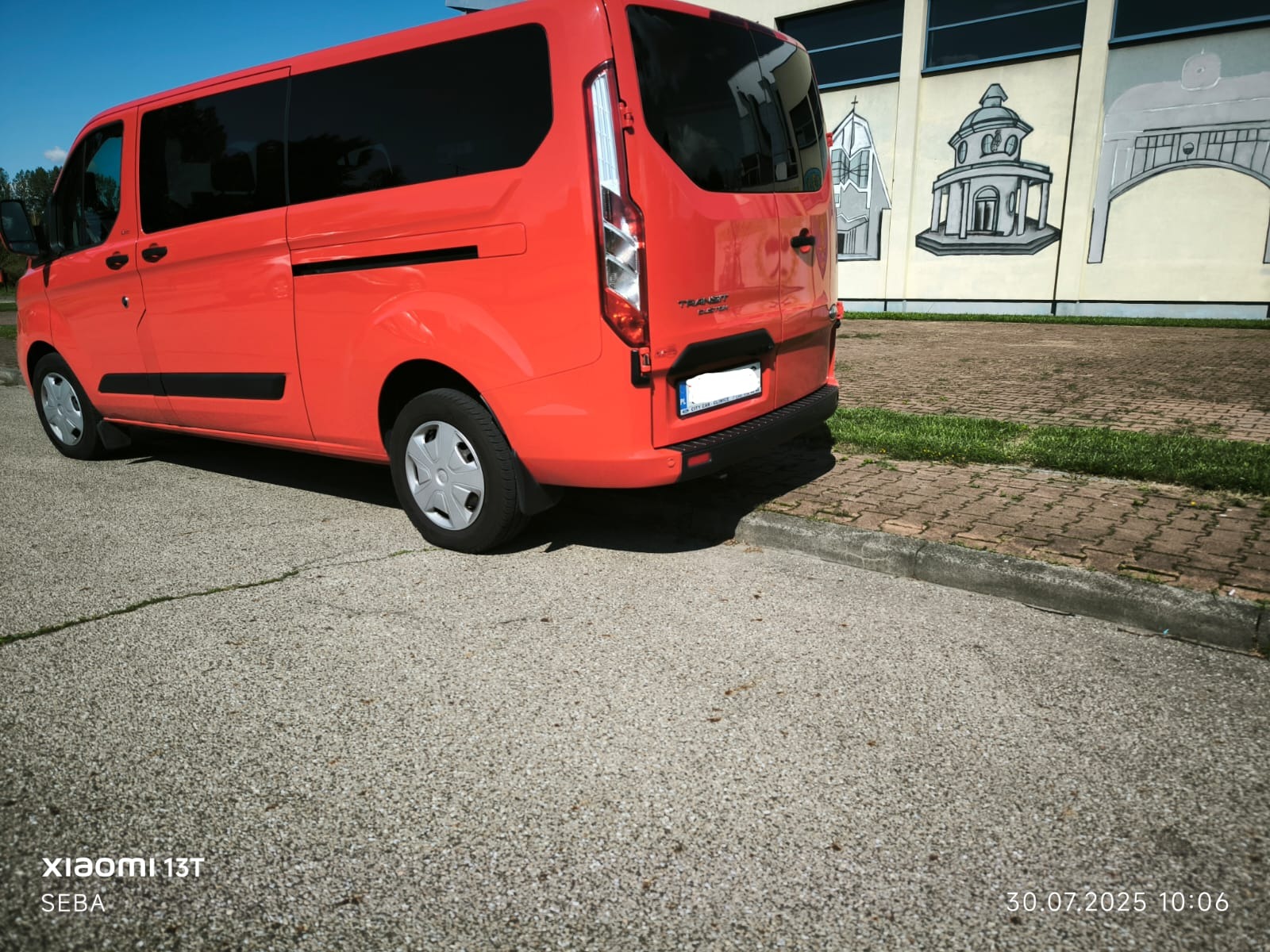Pomarańczowy bus Ford Transit z przyciemnianymi szybami, zaparkowany na zewnątrz, z widocznym fragmentem budynku z malowidłami na ścianie. Tył pojazdu.