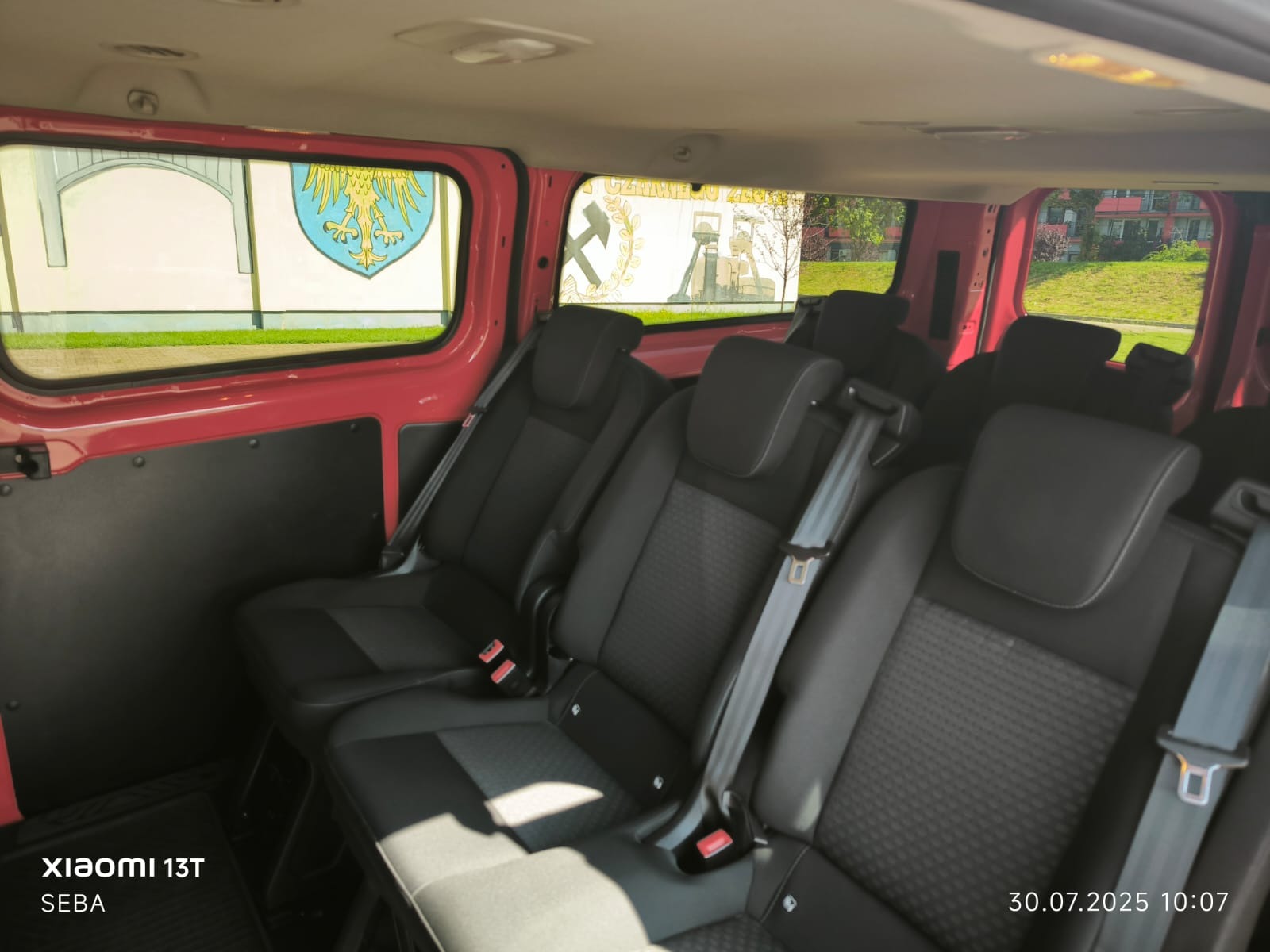 Wnętrze czerwonego busa z trzema rzędami czarnych foteli. Widok z tyłu pojazdu, przez okna widać budynki i murale.