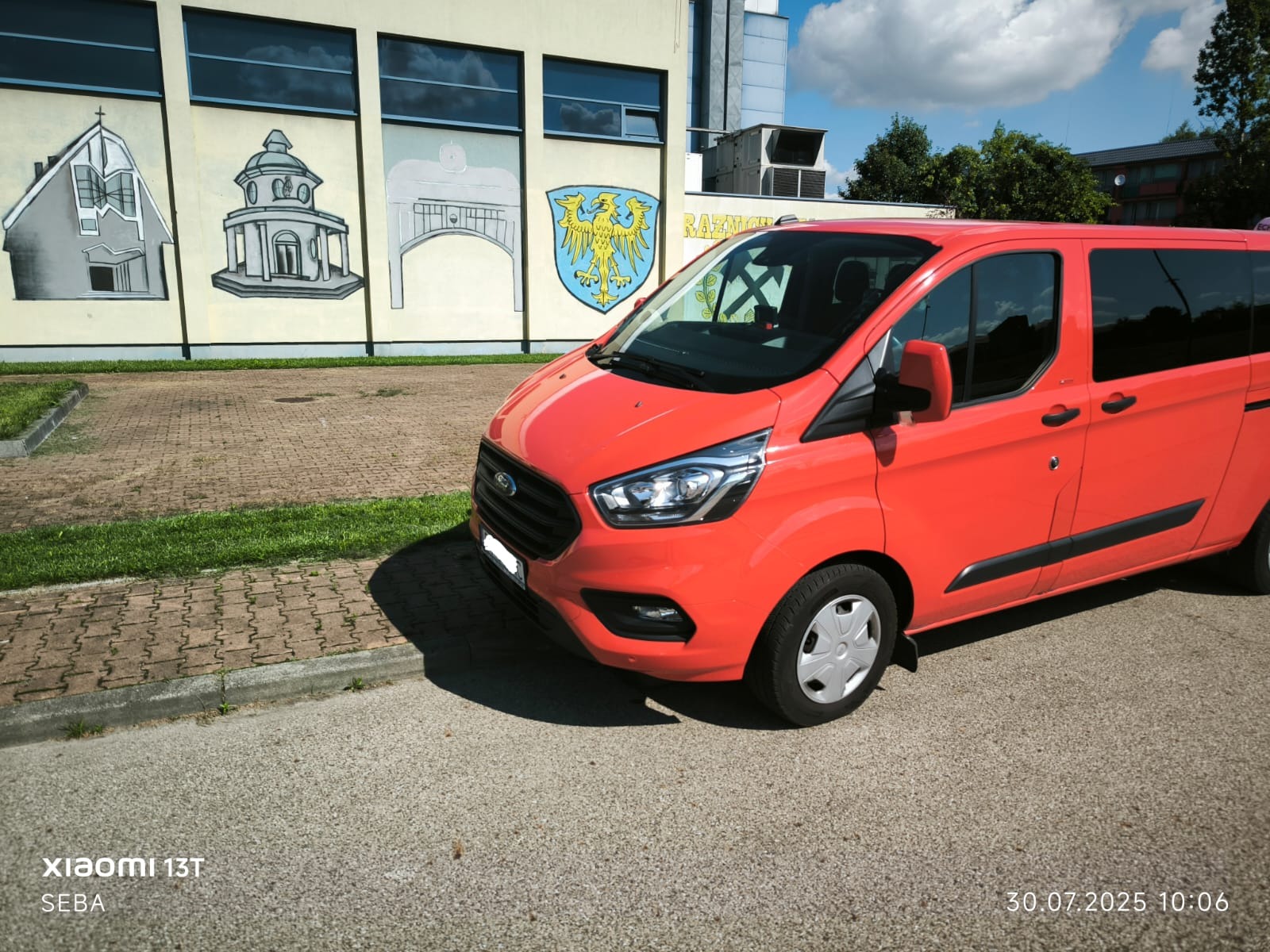 Czerwony bus Ford Transit na tle budynku z muralami. Pojazd zaparkowany na parkingu, widoczne detale karoserii i opon. Słoneczny dzień.