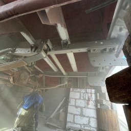 MetalClean Jakub Herdzik - Pracownik w kombinezonie, w zapylonym warsztacie, czyści podwozie pojazdu. Widoczne elementy konstrukcyjne i zawieszenie pojazdu w trakcie obróbki.