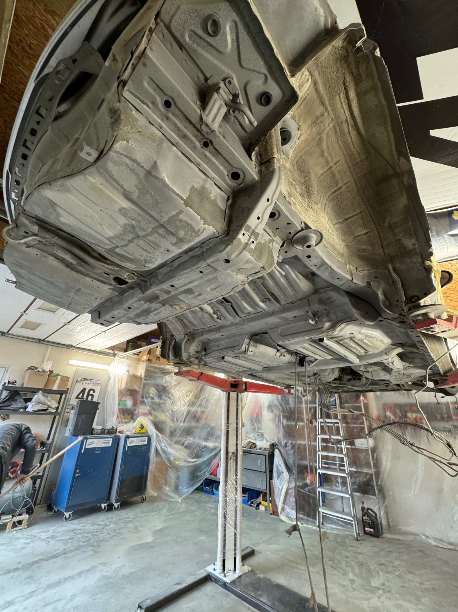 Podwozie samochodu na podnośniku w warsztacie, widok od dołu. Widoczne elementy konstrukcyjne i zabezpieczenia antykorozyjne. W tle narzędzia i wyposażenie warsztatowe.