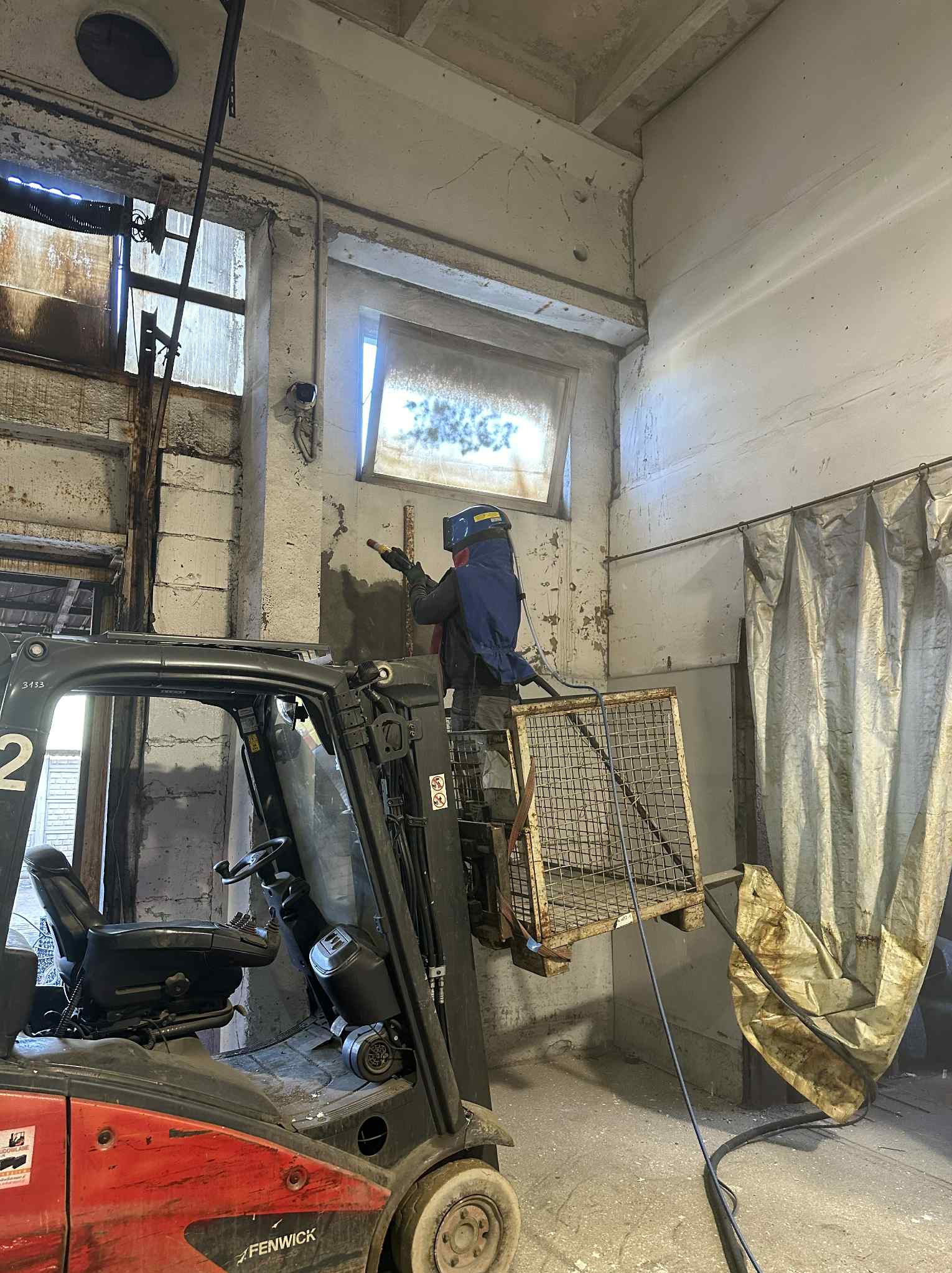 Pracownik w masce ochronnej, na podnośniku widłowym, czyści ścianę w hali przemysłowej. Widoczne ślady po czyszczeniu i okno z zasłoną.