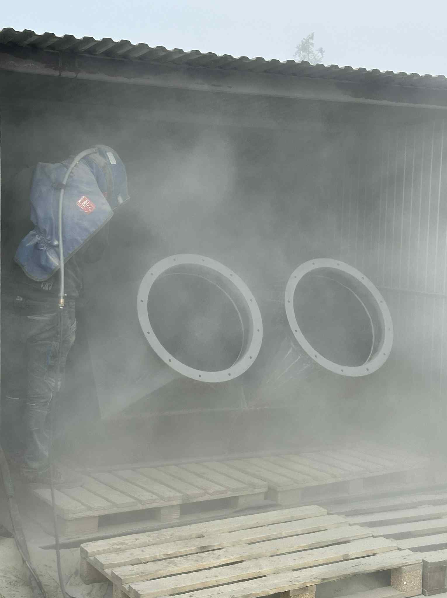 Pracownik w kombinezonie ochronnym czyści metalową konstrukcję z dwoma okrągłymi otworami, używając specjalistycznego sprzętu w zamkniętym pomieszczeniu.