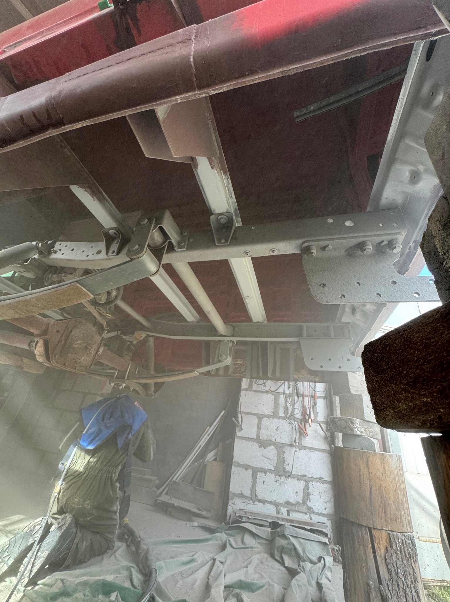 Pracownik w kombinezonie, w zapylonym warsztacie, czyści podwozie pojazdu. Widoczne elementy konstrukcyjne i zawieszenie pojazdu w trakcie obróbki.