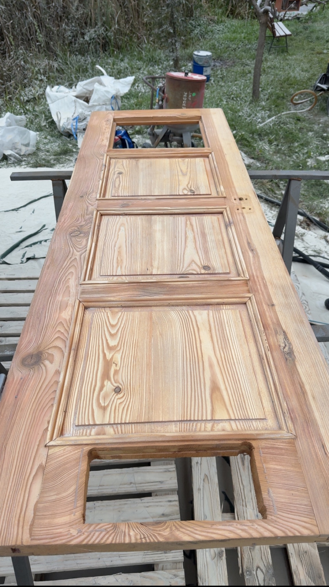 Drewniane drzwi po renowacji, leżące na metalowym stojaku. Widoczne słoje drewna i detale rzeźbienia. W tle sprzęt do piaskowania i otoczenie pokryte białym nalotem.