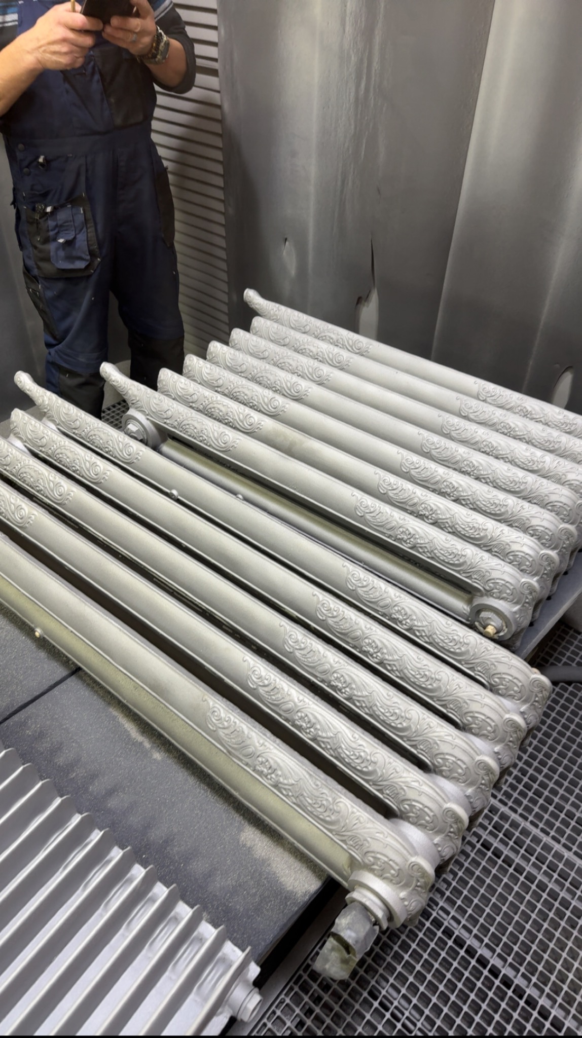 Rzędy odnowionych, dekoracyjnych grzejników w odcieniach szarości, ułożonych na metalowej kratownicy w warsztacie. Osoba w kombinezonie robi zdjęcie telefonem.