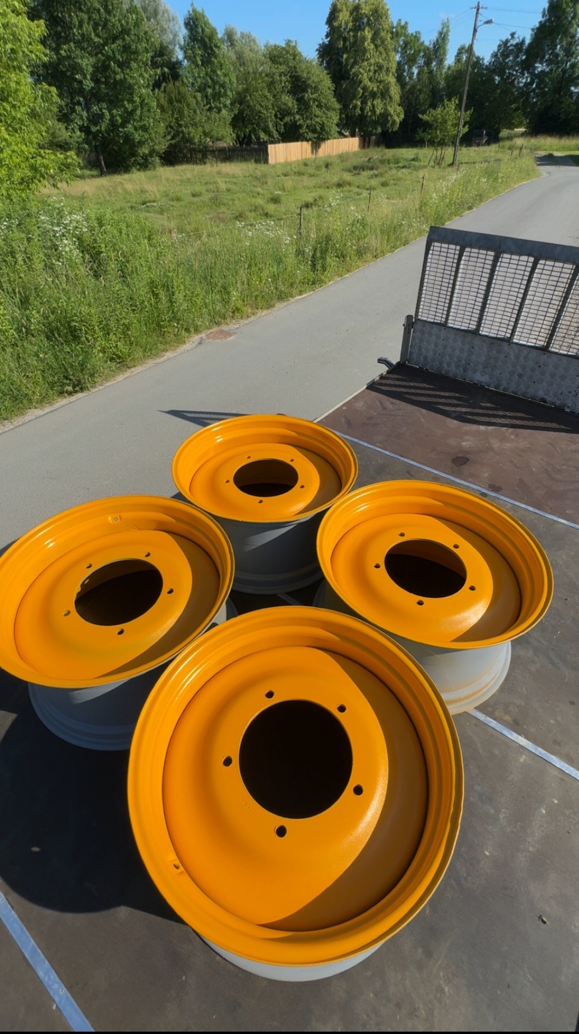 Cztery pomarańczowe felgi po malowaniu proszkowym, ułożone na przyczepie w słoneczny dzień. Widoczne otwory montażowe i szary spód felg. Tło stanowi zielona łąka i droga.