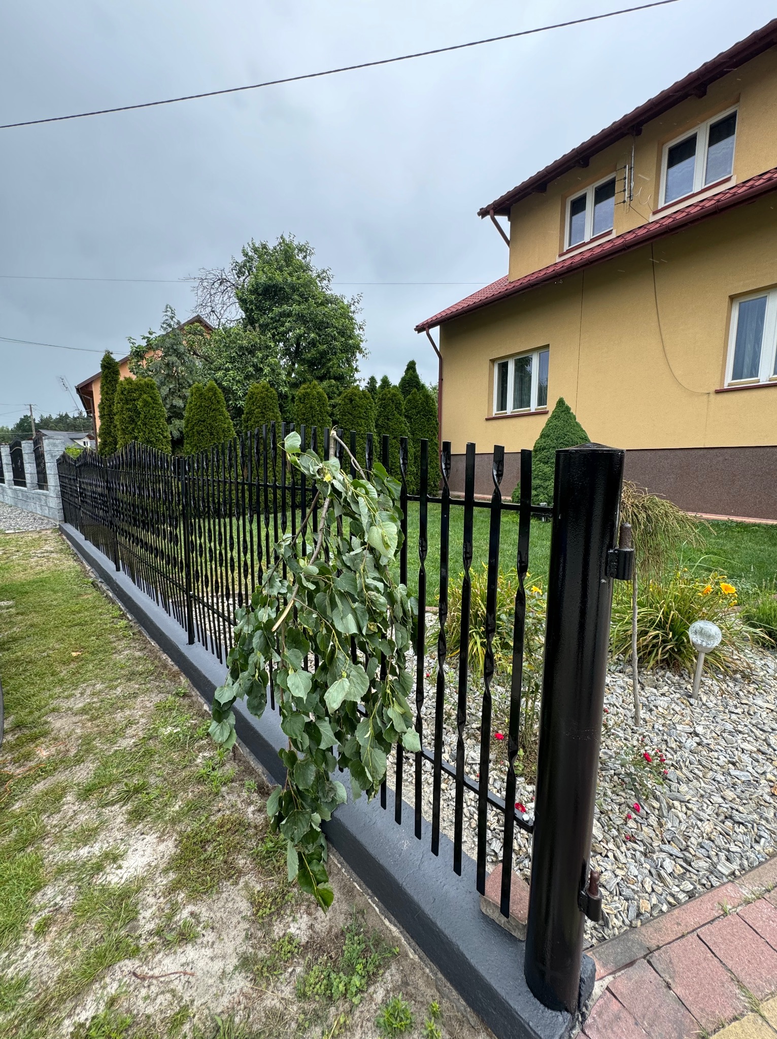 Czarny, metalowy płot z ozdobnymi prętami na tle domu z ogrodem. Na płocie zwisają gałęzie drzewa. Widoczne kamienie ozdobne i trawa.