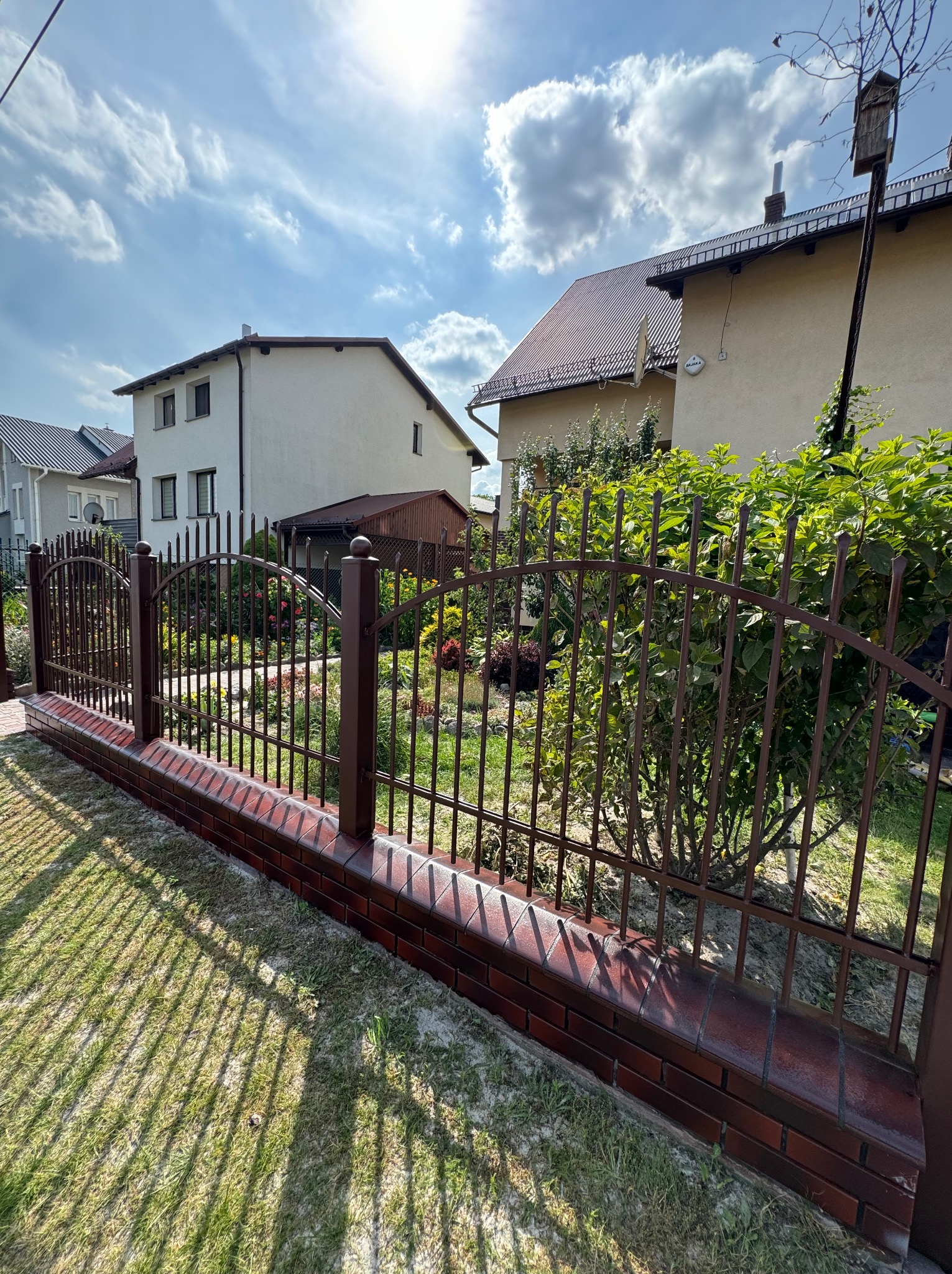 Brązowe ogrodzenie metalowe na ceglanej podmurówce, z widokiem na zadbany ogród i domy w tle, w słoneczny dzień. Cień ogrodzenia na trawie.